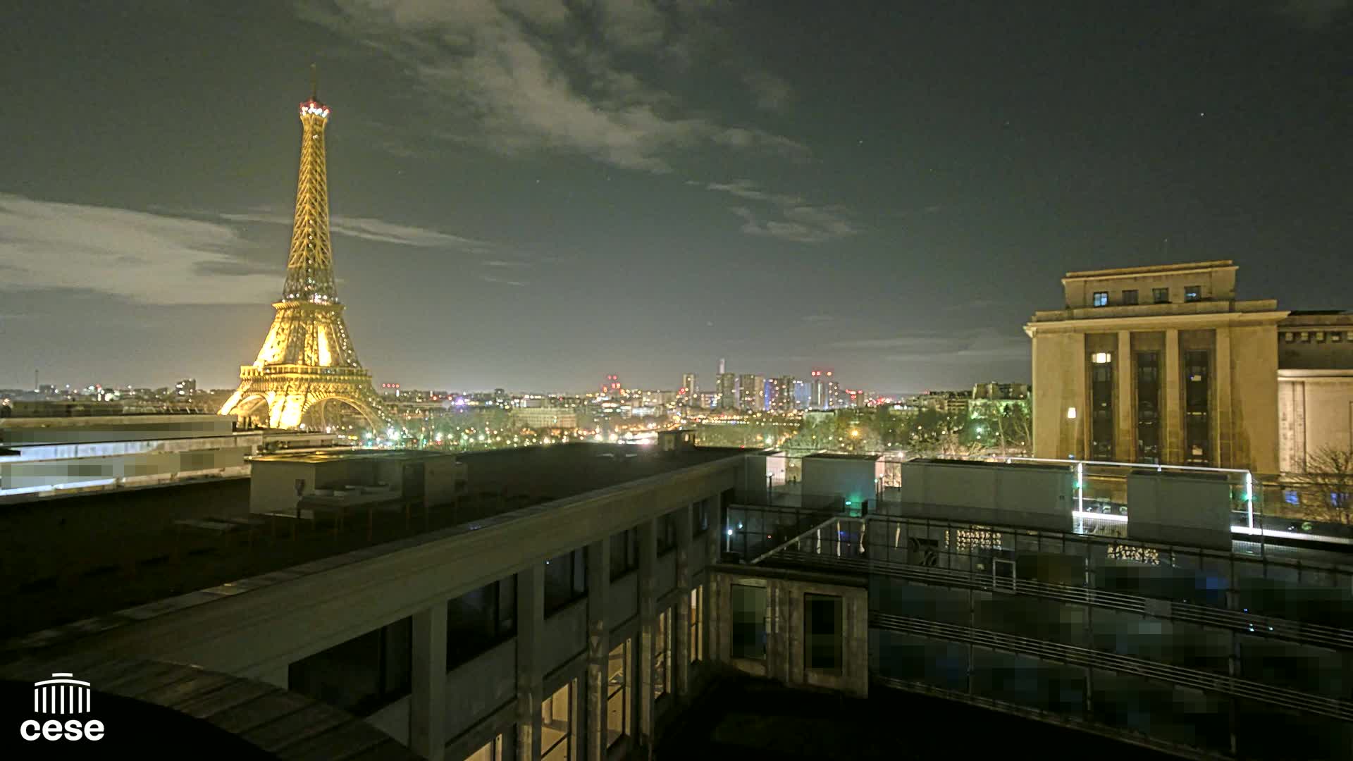 A nighttime view of the Eiffel Tower illuminated, a cityscape, and a large building, under a partly cloudy sky.