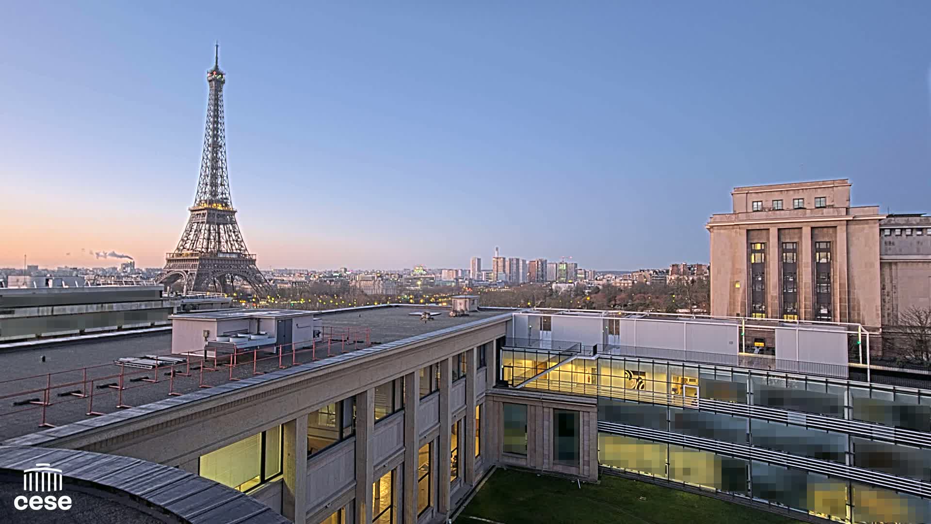 A rooftop view of the Eiffel Tower at dusk, with city buildings and a clear sky in the background.