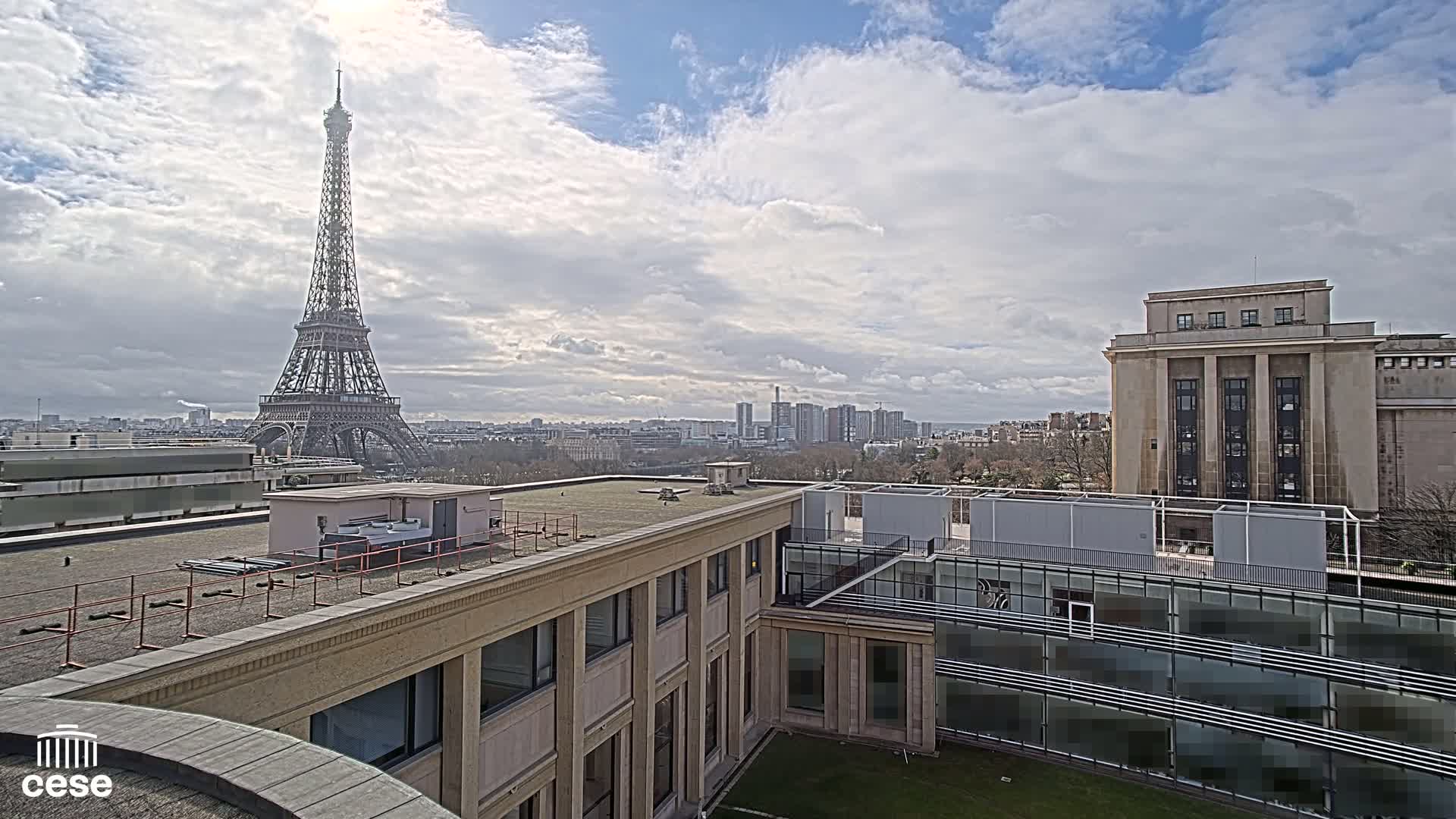 A view from a rooftop shows the Eiffel Tower in the distance under a partly cloudy sky, with city buildings and trees in the foreground.