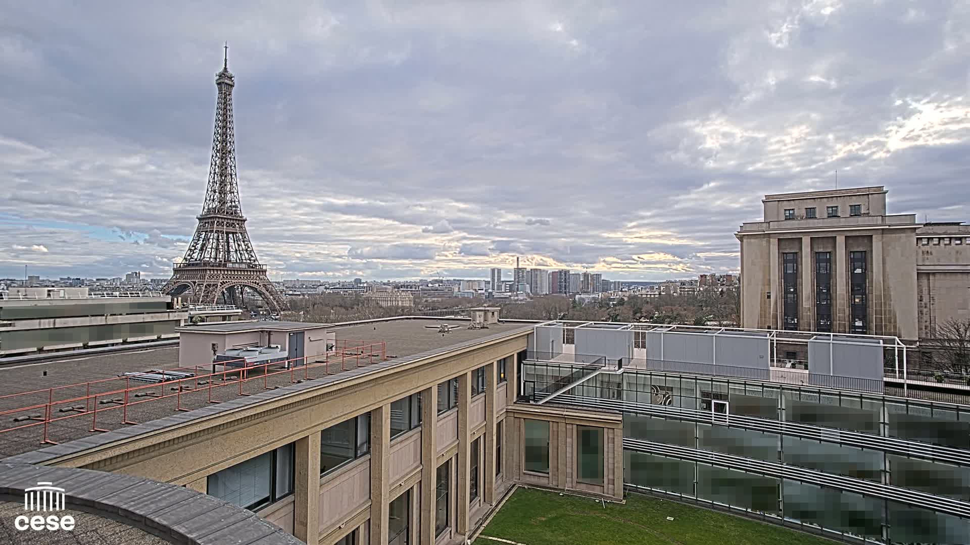 A rooftop view of Parisian architecture, including the Eiffel Tower in the distance, under a cloudy sky.