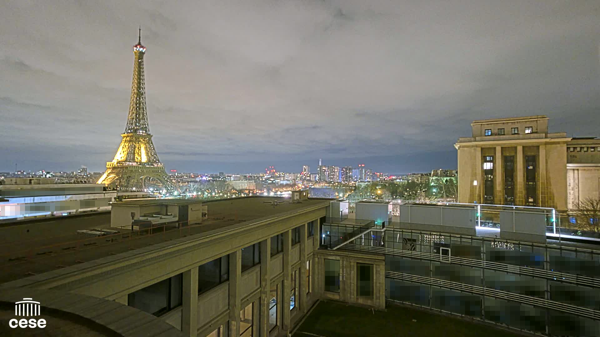 A nighttime view of the Eiffel Tower illuminated against a cloudy sky, overlooking a city skyline and a modern building with a flat roof.