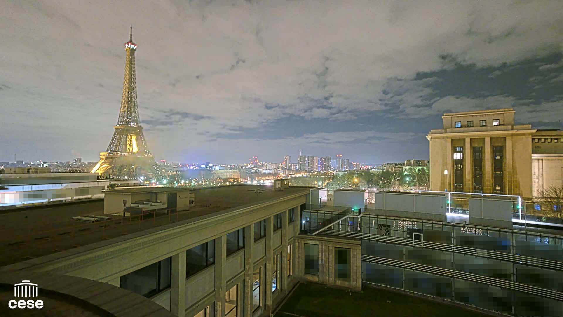 A nighttime view of the Eiffel Tower and a city skyline from a rooftop, under a cloudy sky.