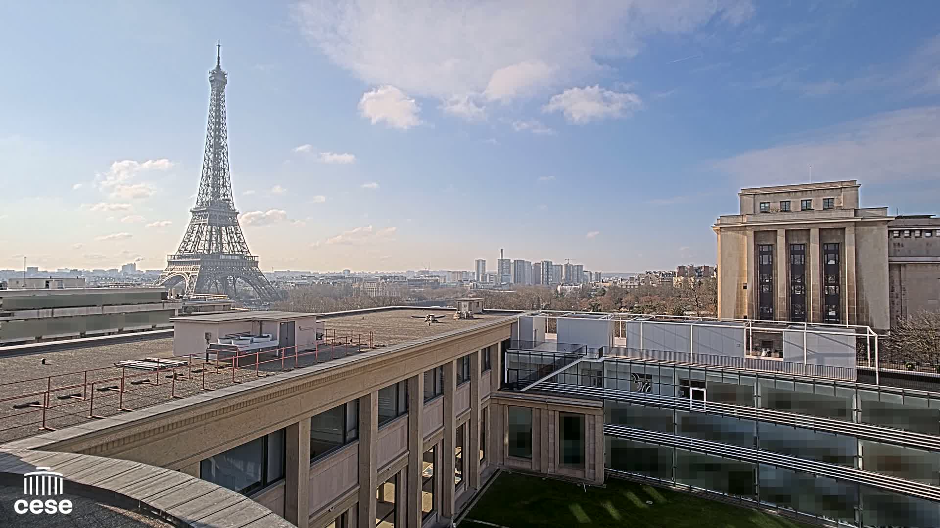 A view from a rooftop shows the Eiffel Tower in the distance under a partly cloudy, sunny sky, with other buildings and a cityscape in the middle ground.