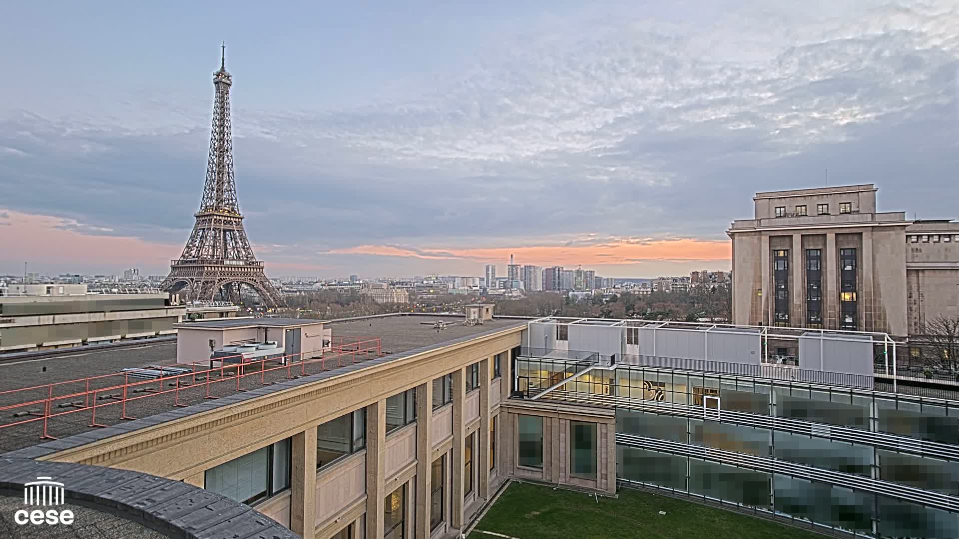 An elevated view of Parisian rooftops at sunset, showcasing the Eiffel Tower in the distance under a partly cloudy sky.