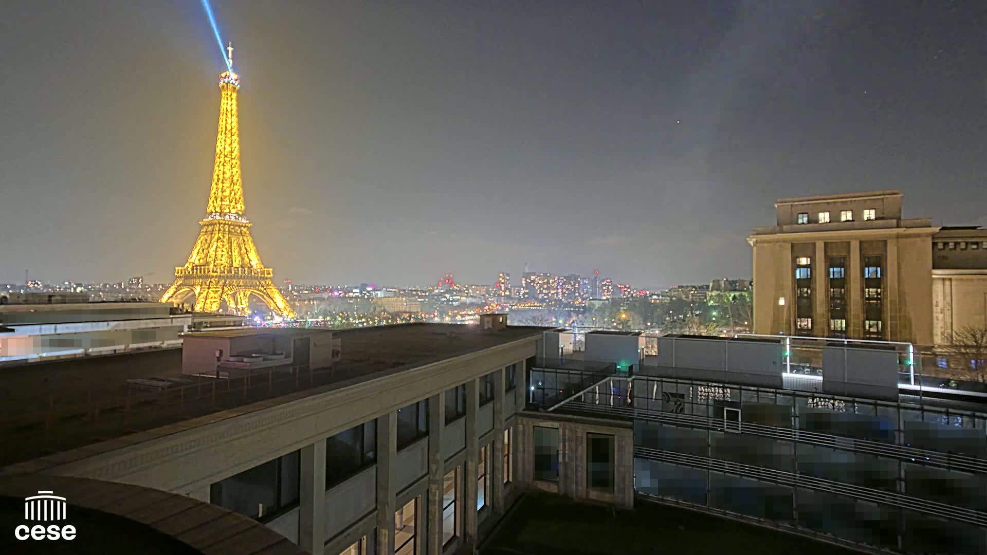 A nighttime view of the Eiffel Tower illuminated in gold, with a city skyline in the background and a building in the foreground under a clear sky.
