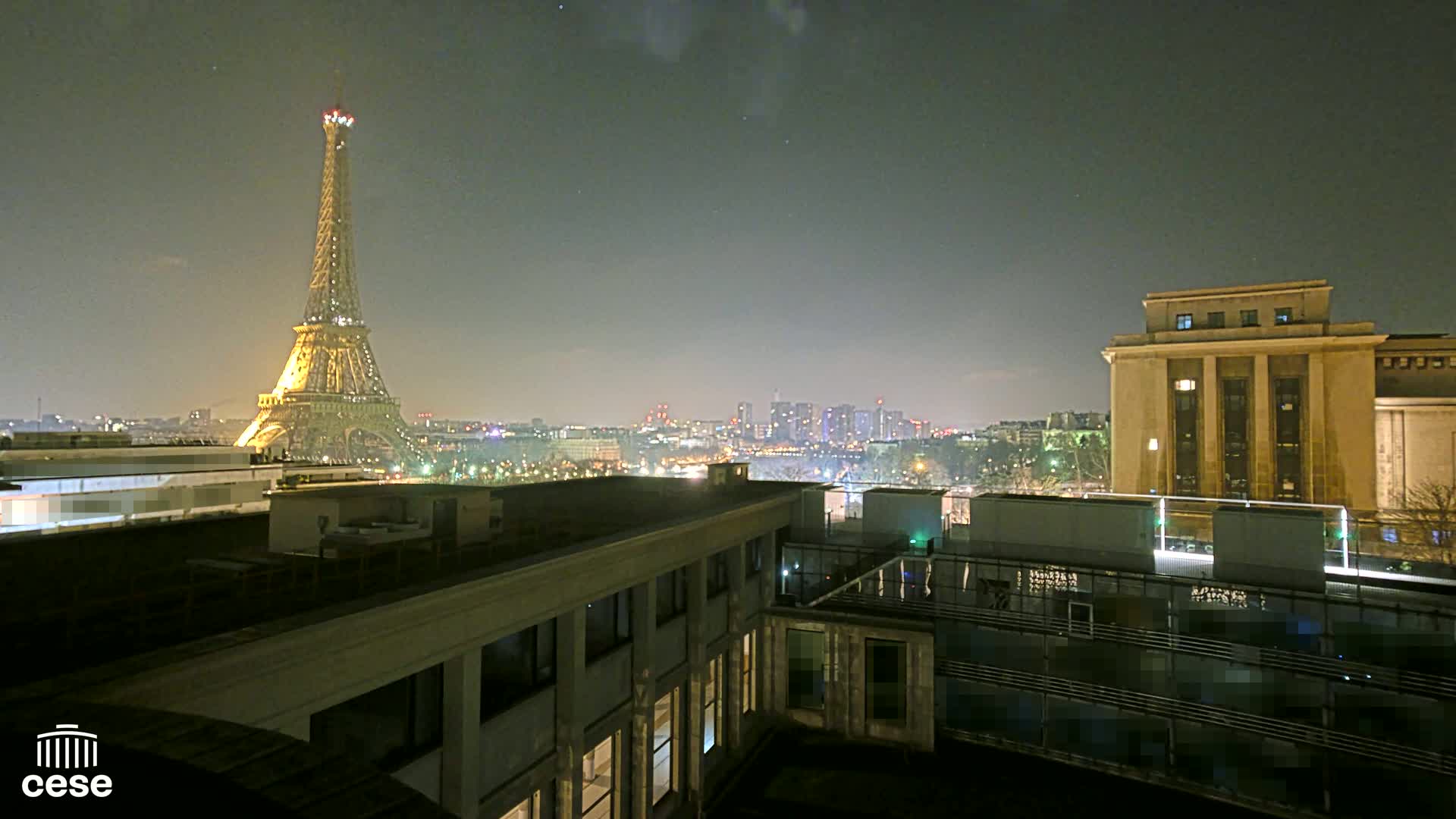 A nighttime cityscape view of the Eiffel Tower and a city skyline, seen from a rooftop with another building in the foreground under a clear, dark sky.