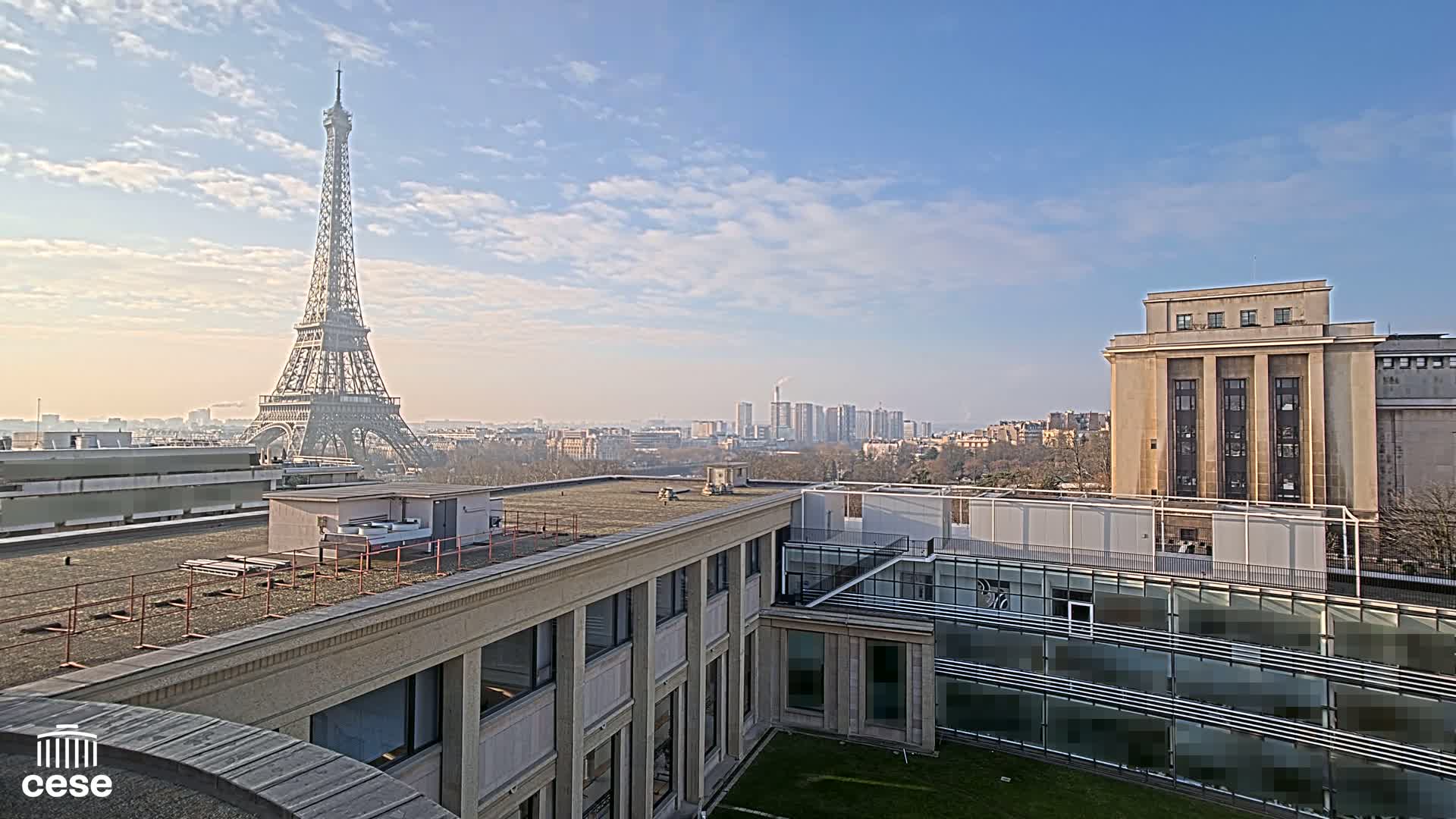 A rooftop view of Paris on a partly cloudy day shows the Eiffel Tower in the distance and a cityscape including several buildings.