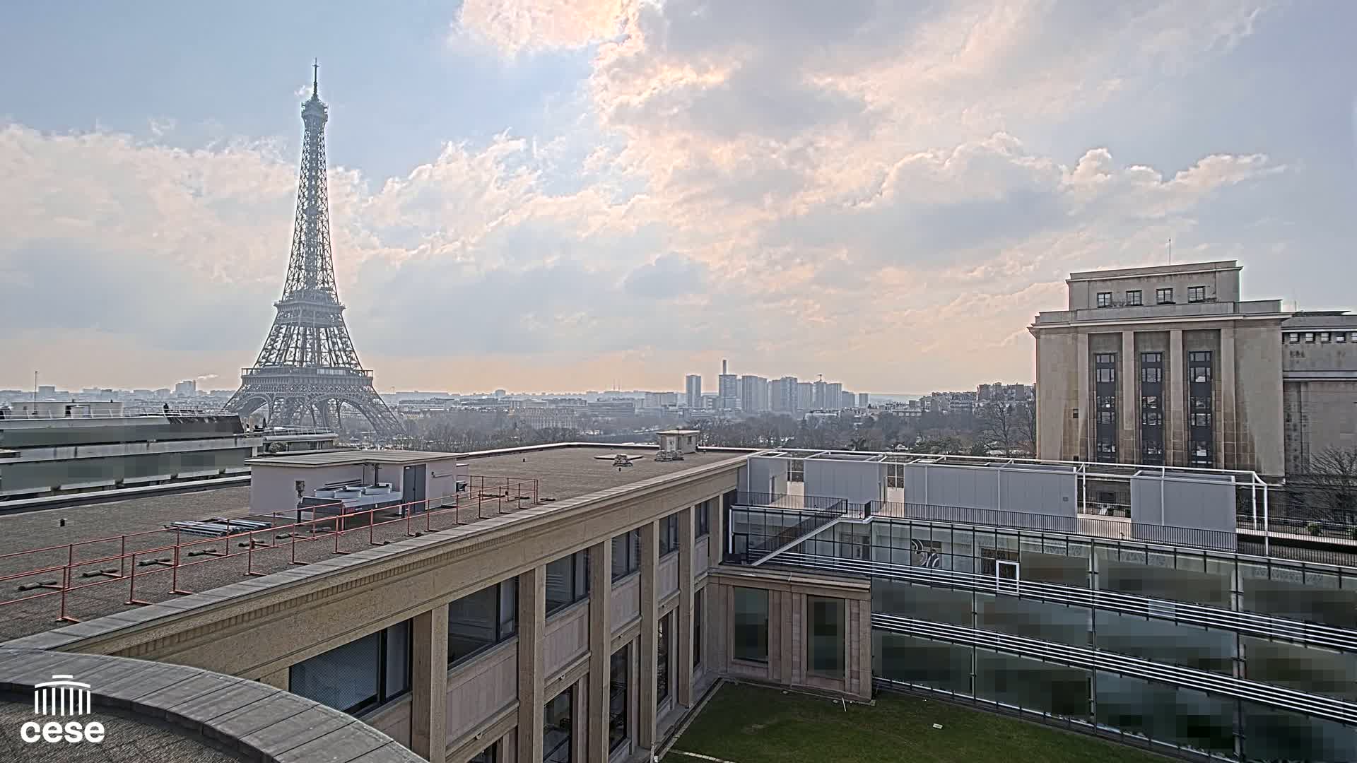 A view from a rooftop shows the Eiffel Tower in the distance under a partly cloudy sky, with other buildings and a cityscape in the background.