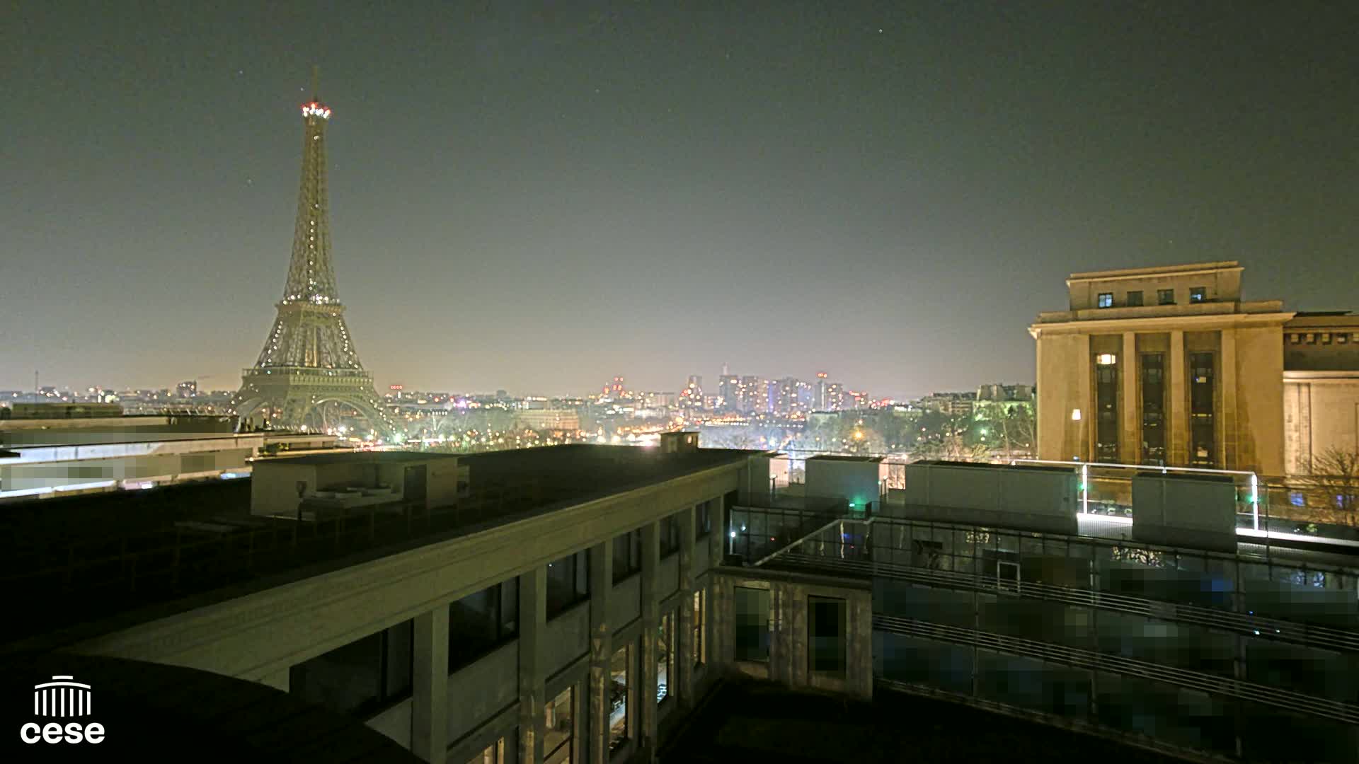 A nighttime view of the Eiffel Tower and a cityscape, seen from a rooftop with another building in the foreground under a clear, dark sky.