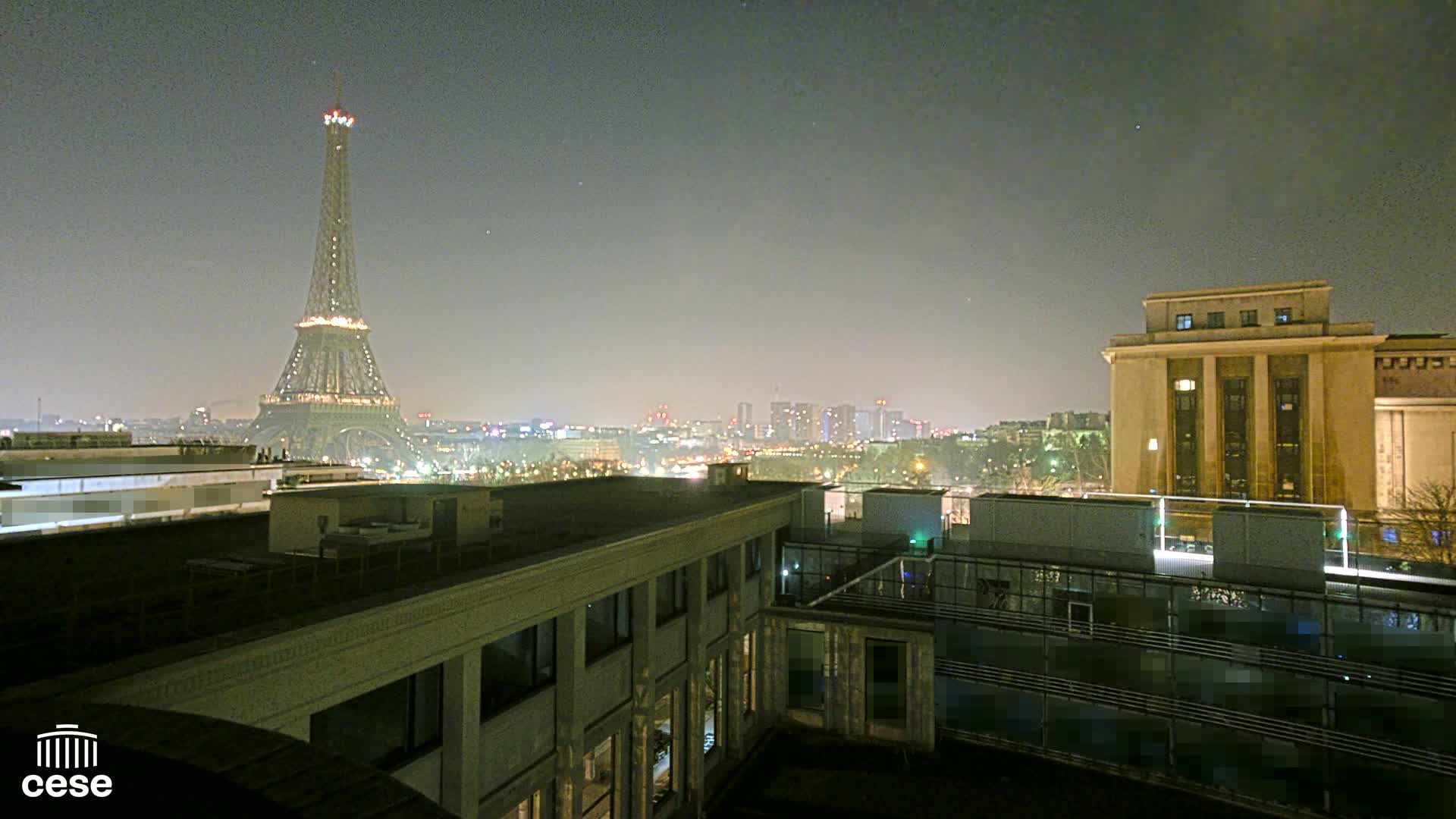 A nighttime view of the Eiffel Tower and a city skyline, seen from a rooftop with a large building in the foreground under a hazy sky.