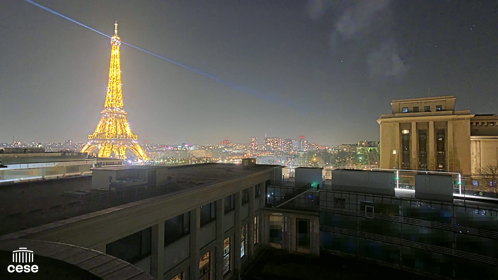 A nighttime view of the Eiffel Tower illuminated, with a city skyline and a large building in the foreground under a clear, dark sky.