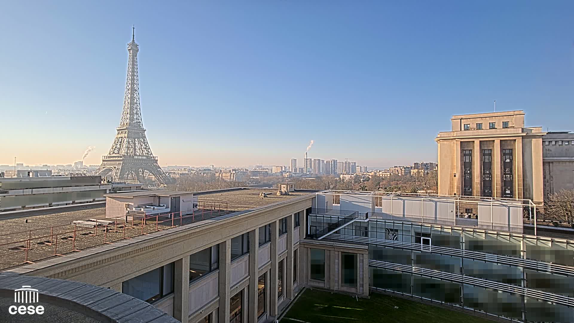 A rooftop view of Paris on a clear, sunny day shows the Eiffel Tower in the distance and a cityscape beyond a modern building.