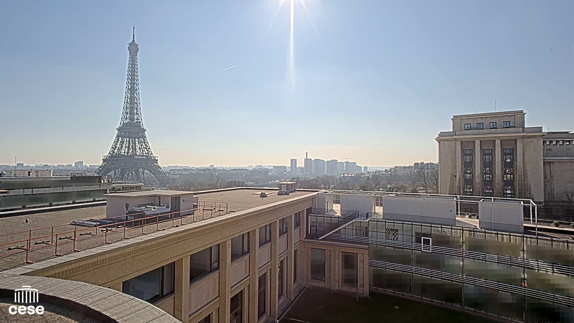 A sunny Parisian rooftop view shows the Eiffel Tower in the distance, a city skyline, and a large tan building.