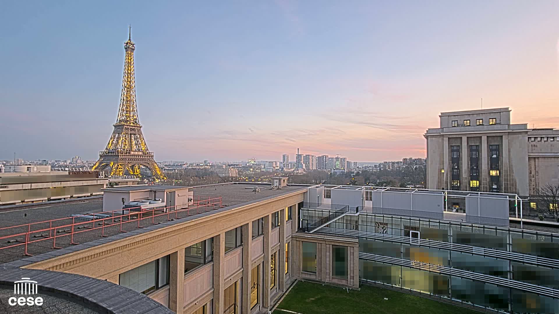 A view from a rooftop shows the Eiffel Tower illuminated at dusk, alongside a cityscape and a large, light-colored building.