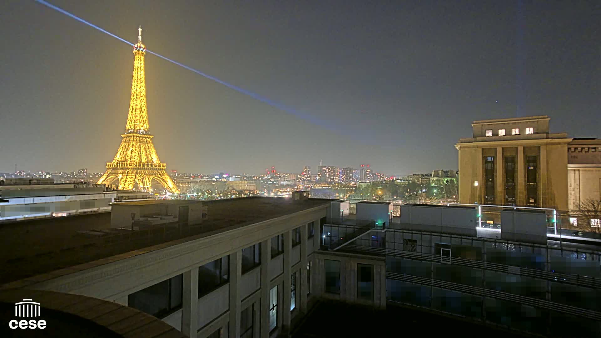 A nighttime view of the Eiffel Tower illuminated, across a cityscape, from a rooftop overlooking other buildings.