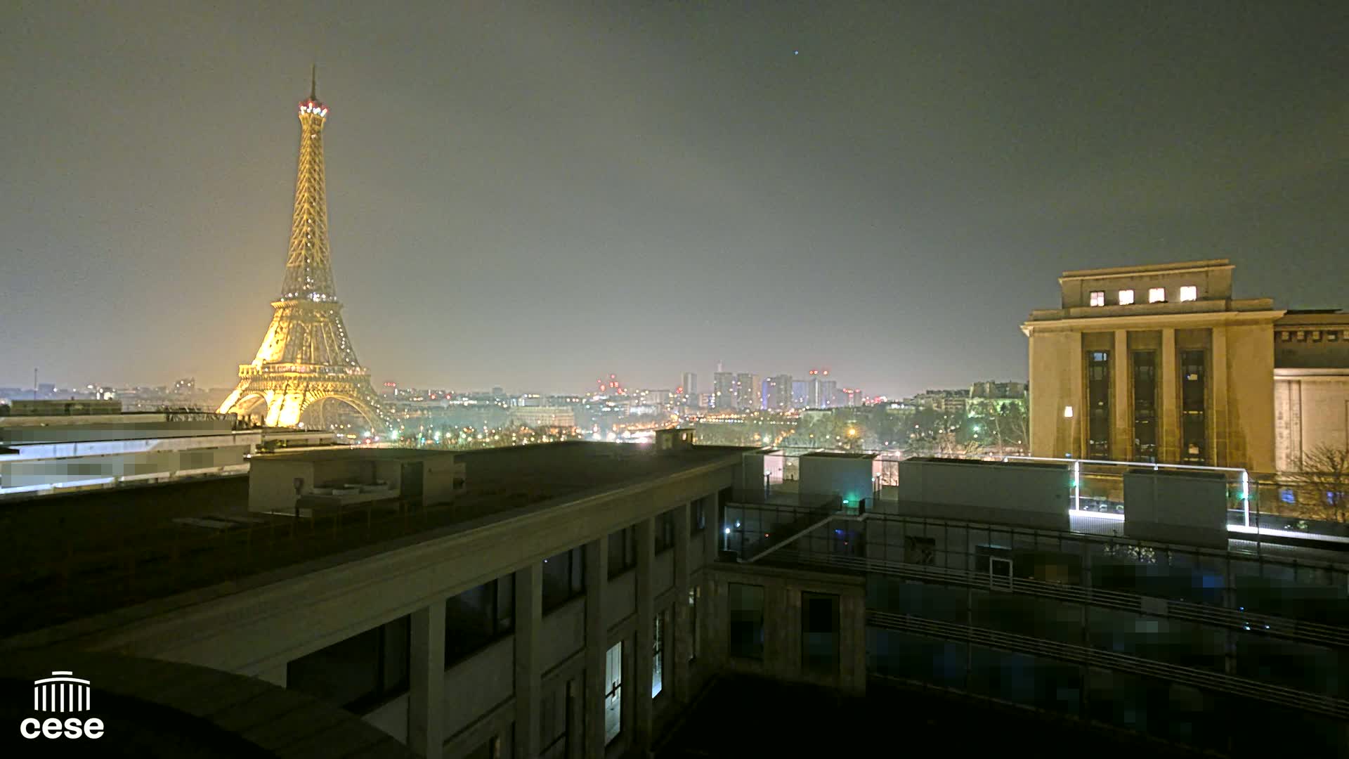 A nighttime view of the Eiffel Tower illuminated, along with a cityscape and a large building, under a hazy sky.