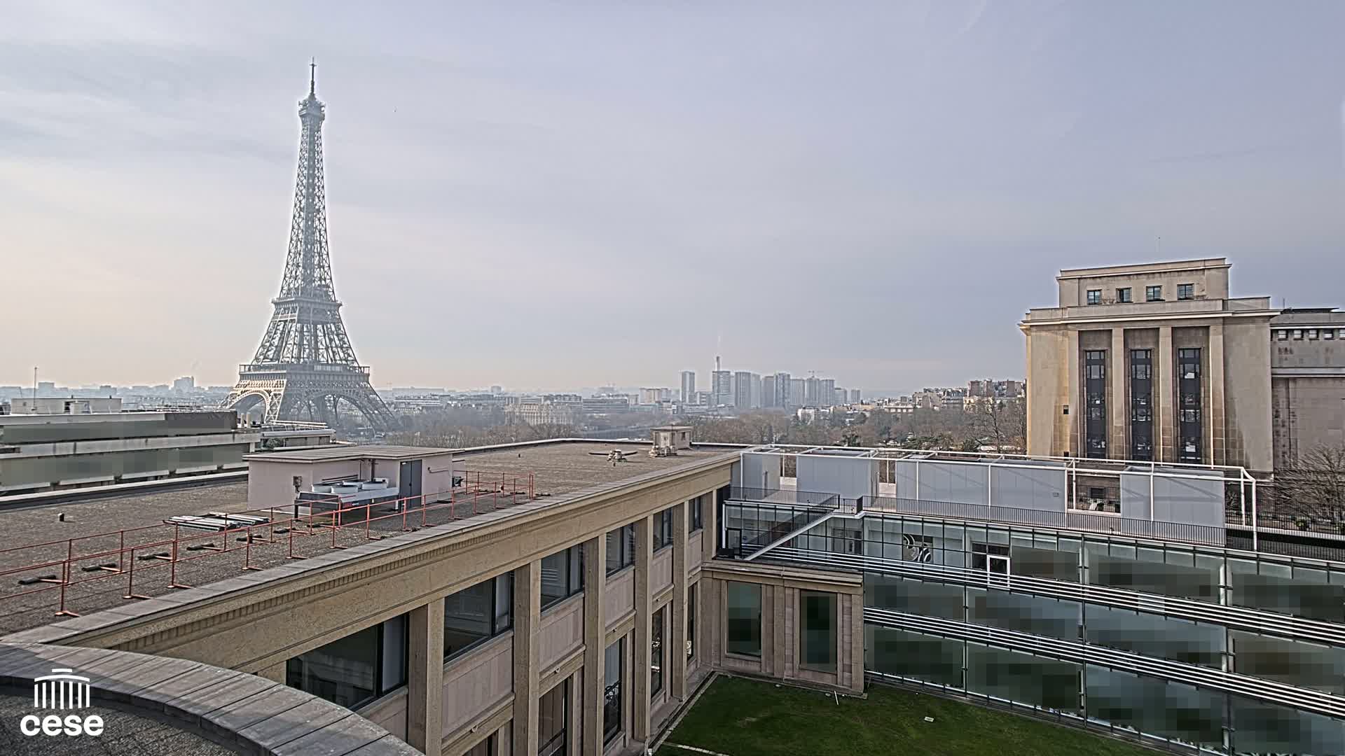 A view from a rooftop shows the Eiffel Tower in the distance on a hazy day, with modern and classical buildings in the foreground.