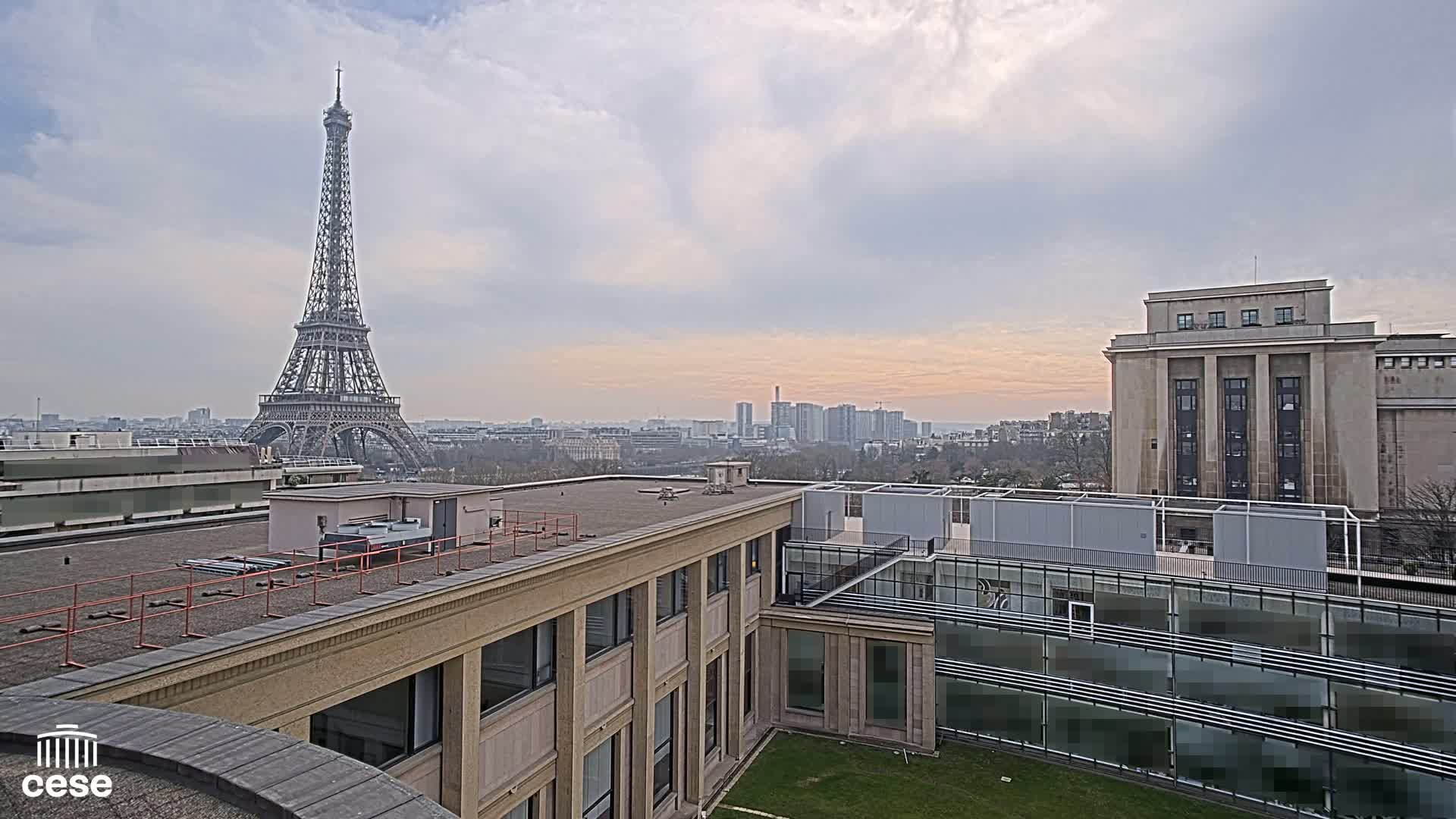 A rooftop view of Paris on an overcast day shows the Eiffel Tower in the distance, city buildings, and a courtyard garden.