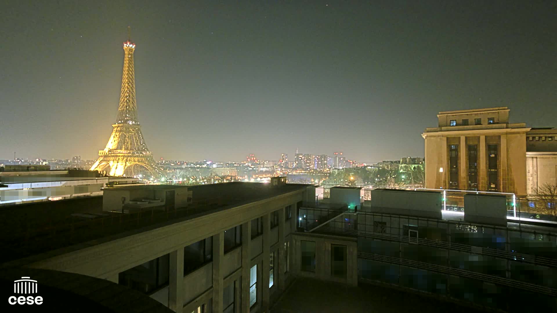 The Eiffel Tower is illuminated at night, overlooking a city skyline and a large building from a rooftop vantage point.