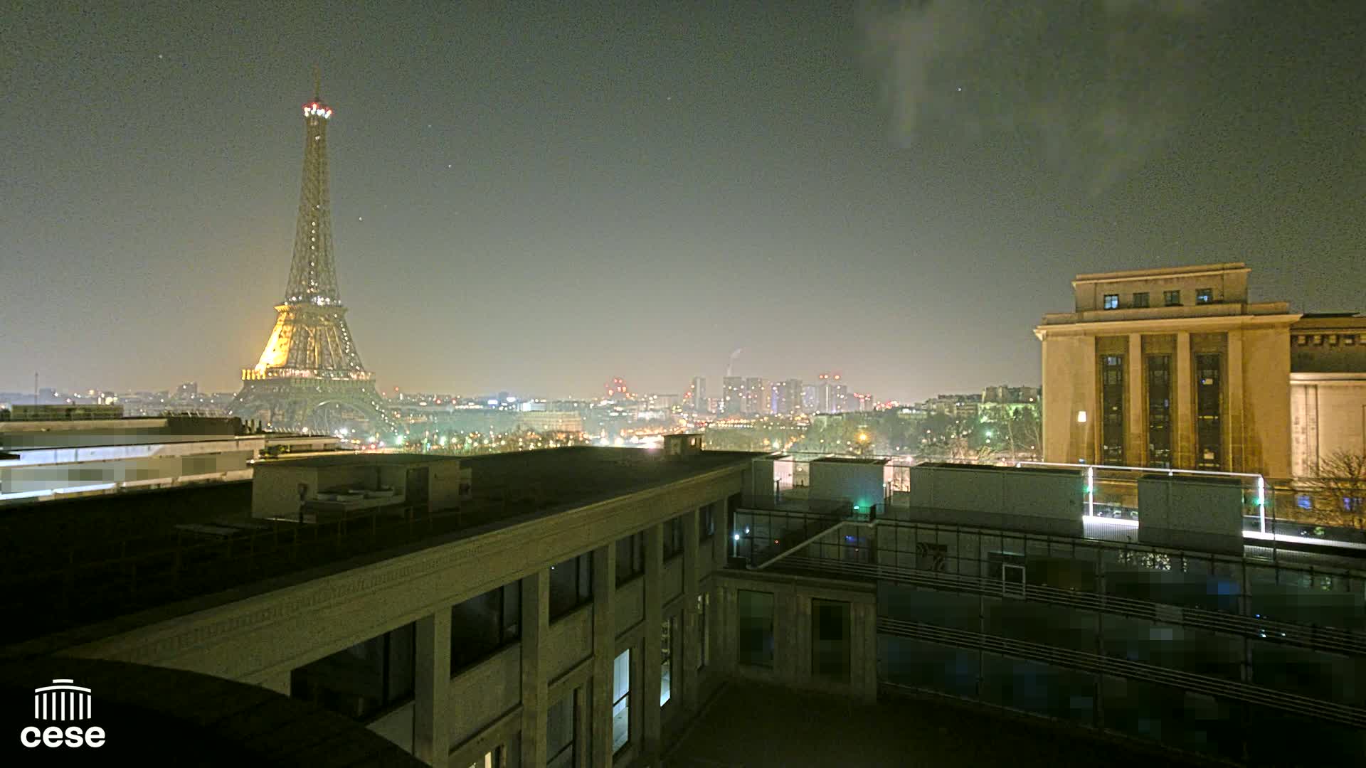 A nighttime view of the Eiffel Tower and Parisian cityscape, including a large building in the foreground, under a hazy sky.