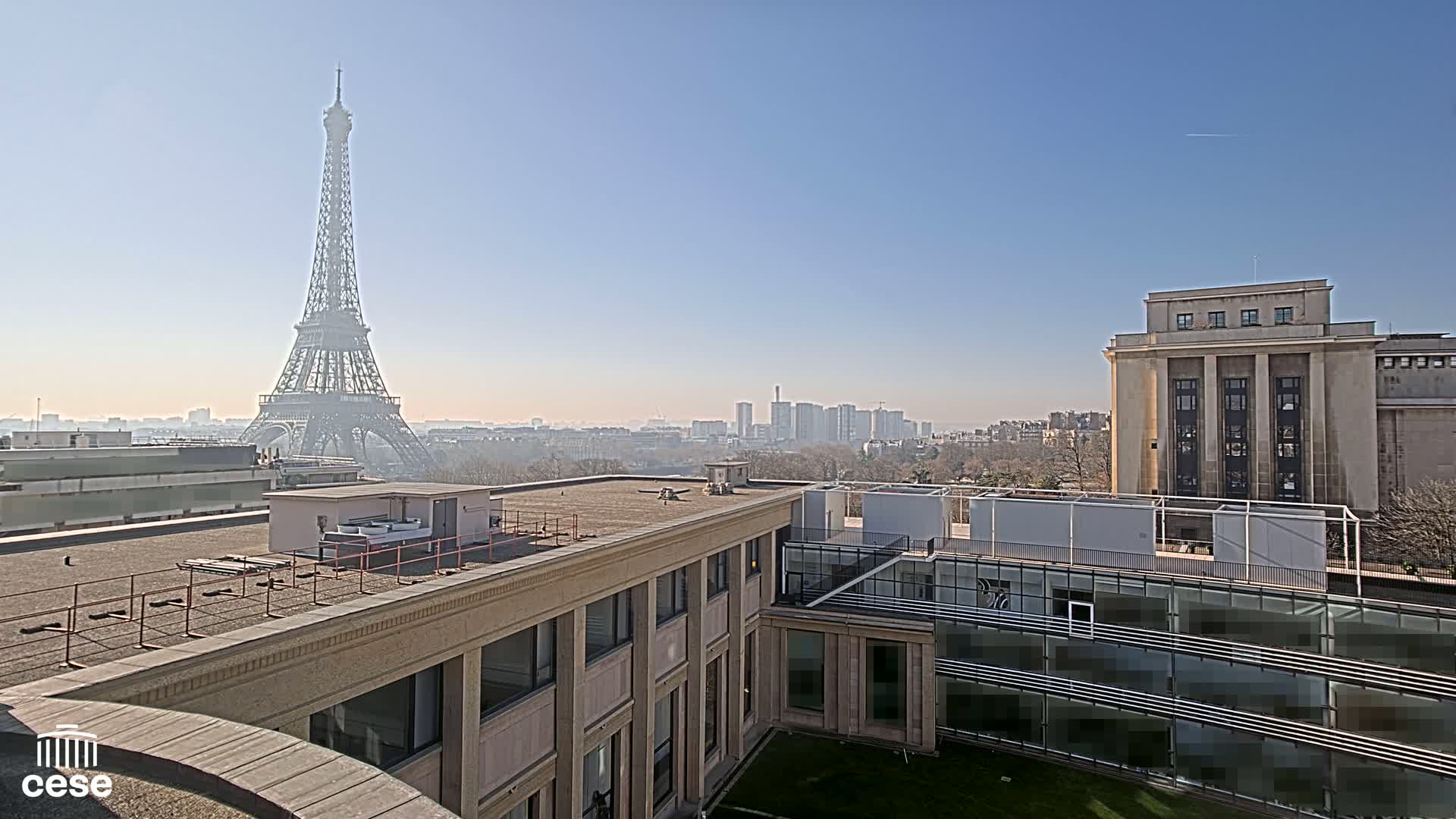 A rooftop view shows the Eiffel Tower in the distance on a clear, sunny day, with modern buildings and a cityscape in the background.