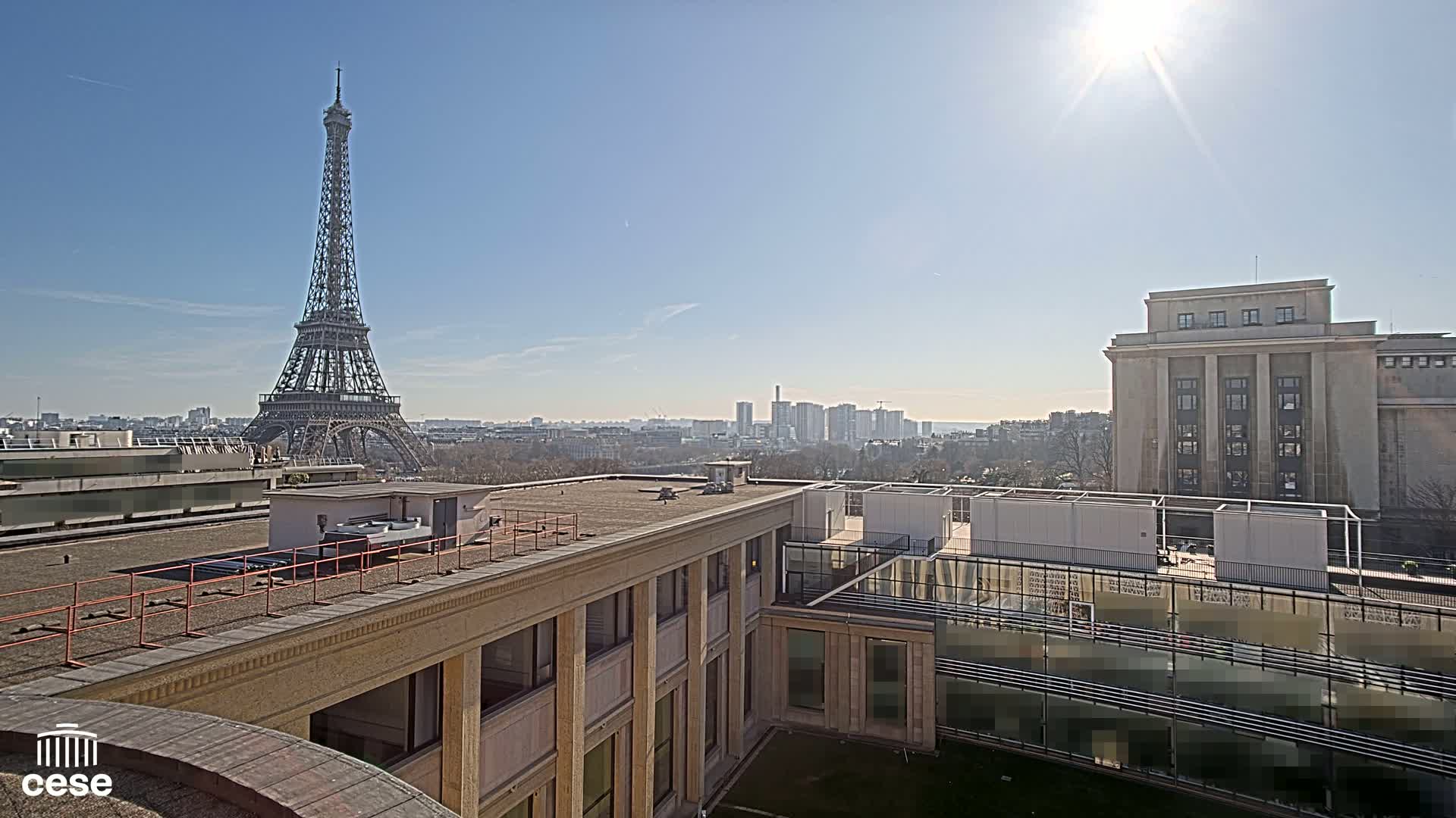 A sunny cityscape view shows the Eiffel Tower in the distance, behind a tan building and a grassy courtyard.
