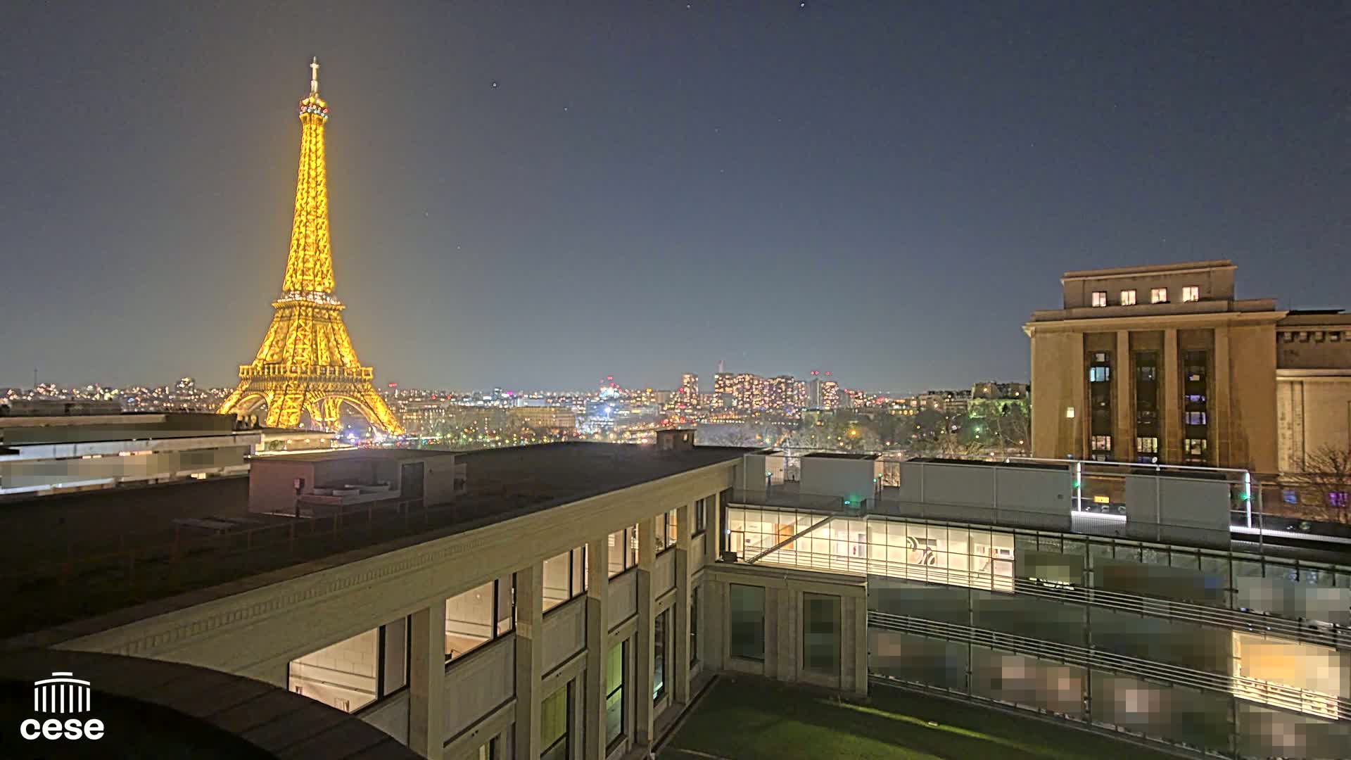A nighttime cityscape shows the Eiffel Tower illuminated, a city skyline in the distance, and modern buildings in the foreground under a clear, dark sky.