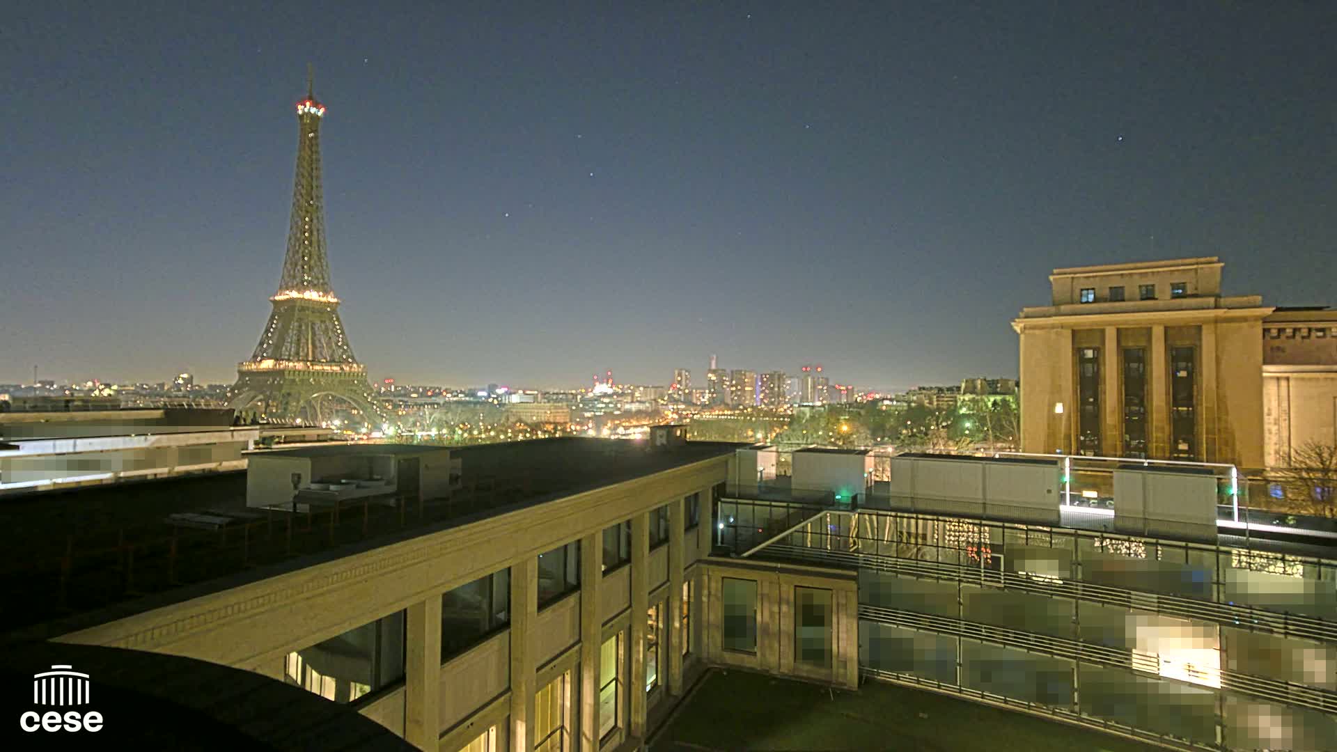 A nighttime view of the Eiffel Tower and a cityscape, seen from a rooftop building with a grassy courtyard.