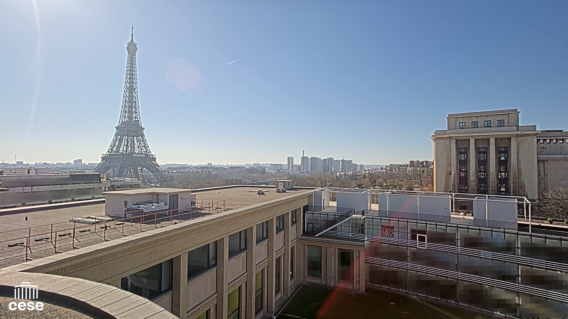 A sunny cityscape view from a rooftop shows the Eiffel Tower in the distance, other buildings, and a grassy courtyard below.