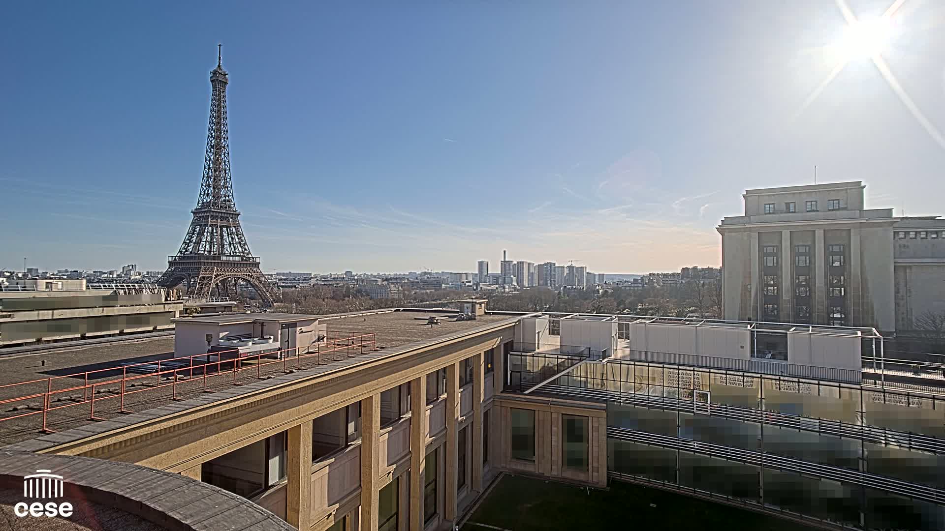 A sunny cityscape view from a rooftop shows the Eiffel Tower in the distance, a large building to the right, and a smaller building in the foreground.