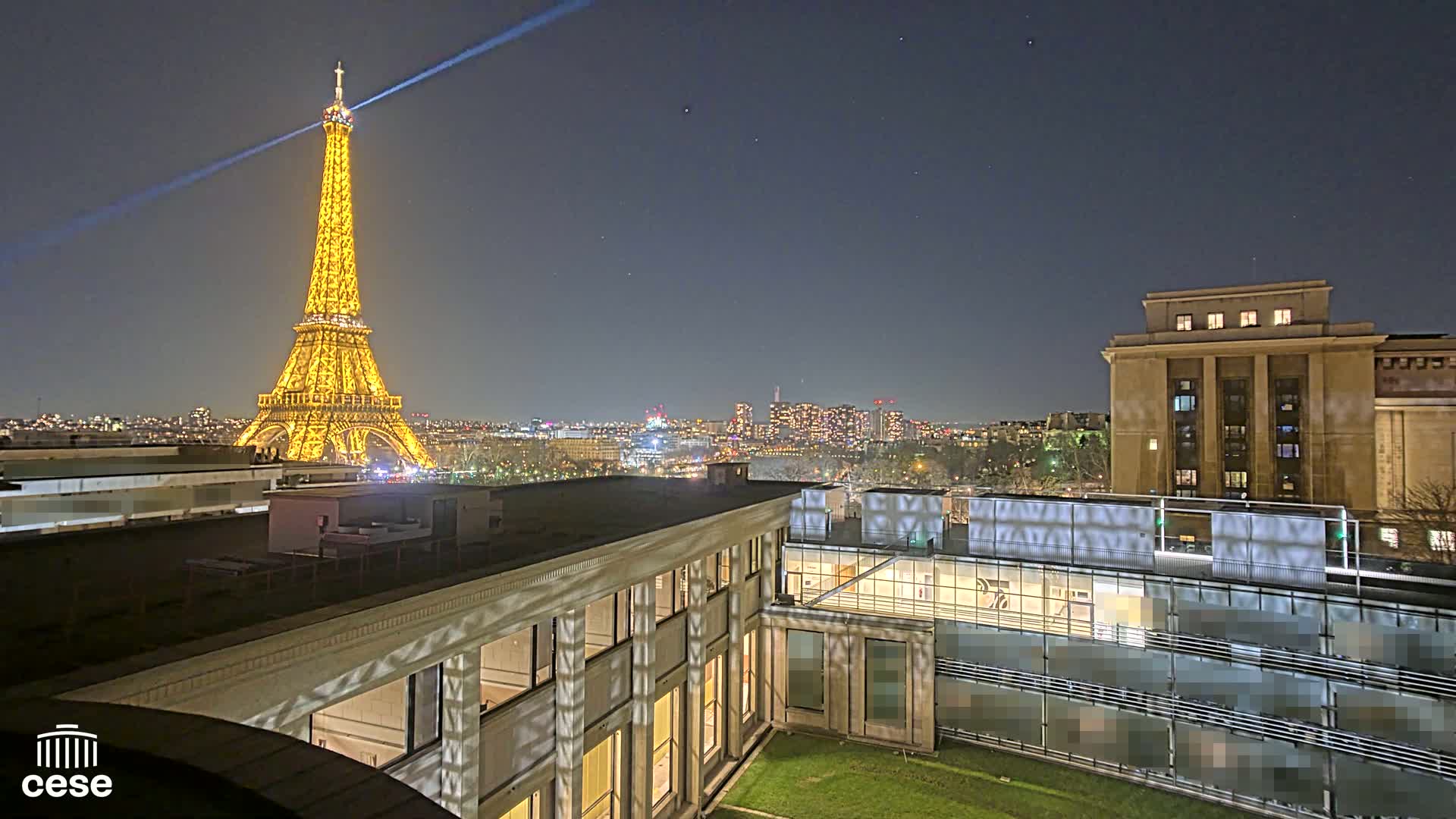 A brightly lit Eiffel Tower is visible at night from a rooftop overlooking a city, with another building and a small grassy area in the foreground.