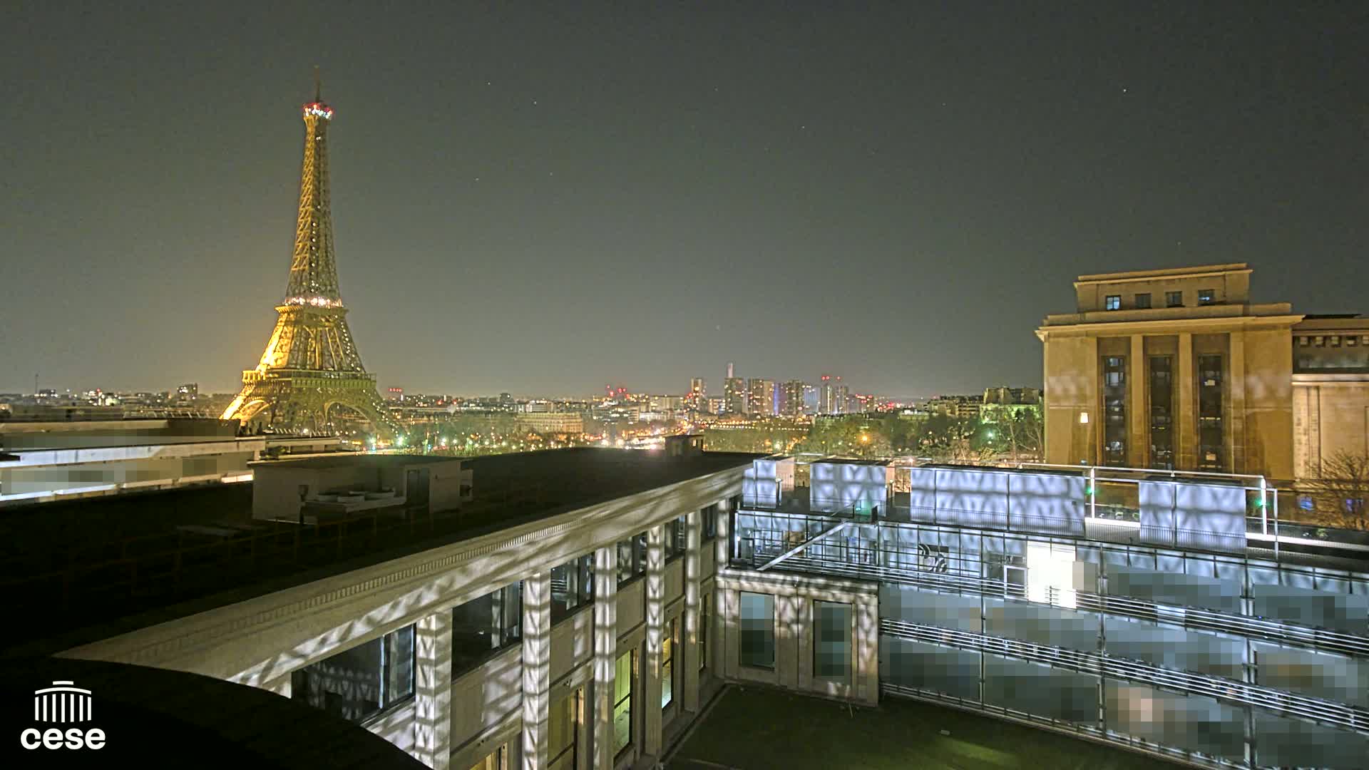 A nighttime view of the Eiffel Tower illuminated against a dark sky, seen from a rooftop overlooking a city skyline and a building with patterned light projections on its walls.