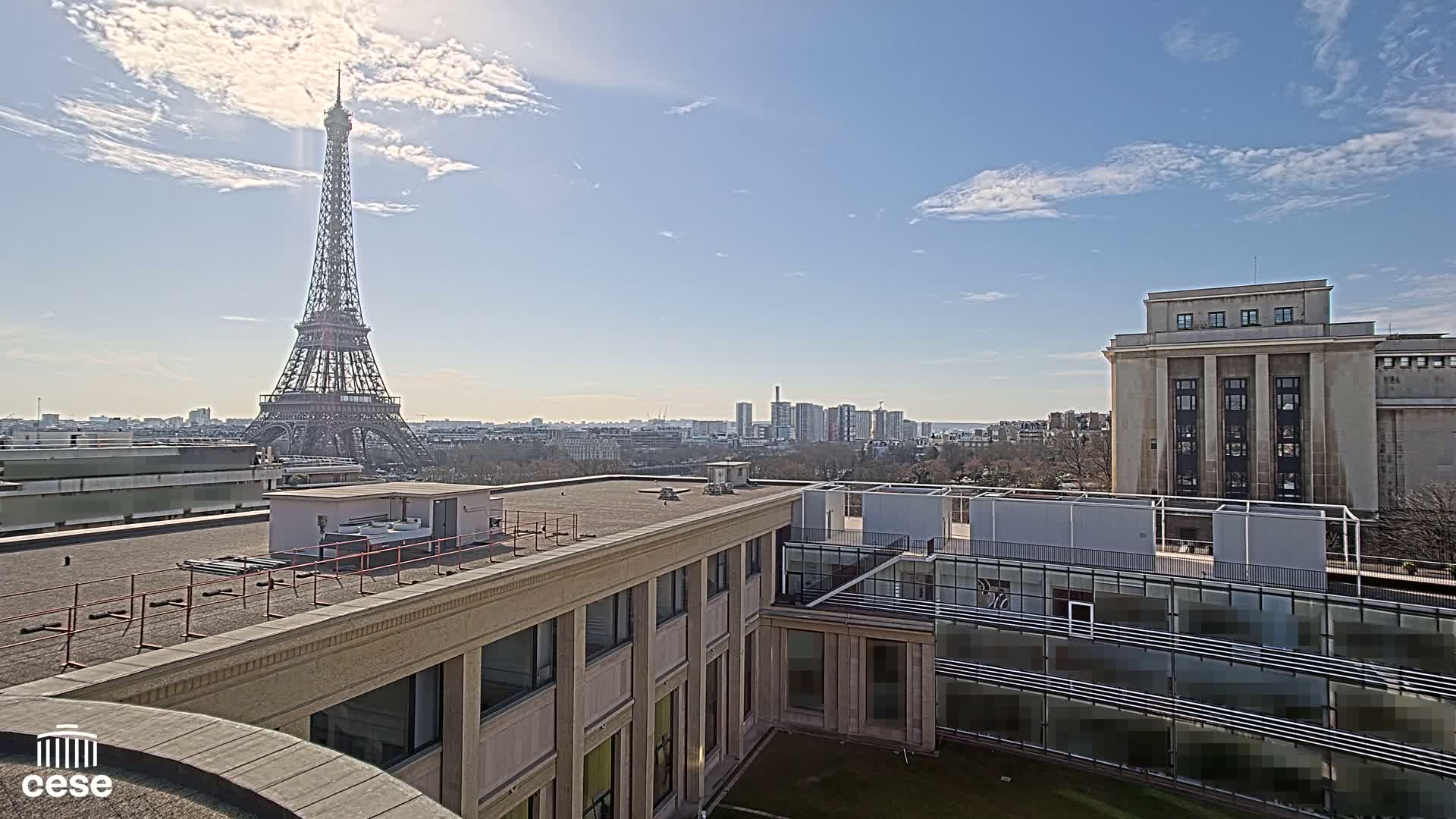 A sunny cityscape view from a rooftop shows the Eiffel Tower in the distance, a building with a flat roof in the foreground, and other buildings and trees.