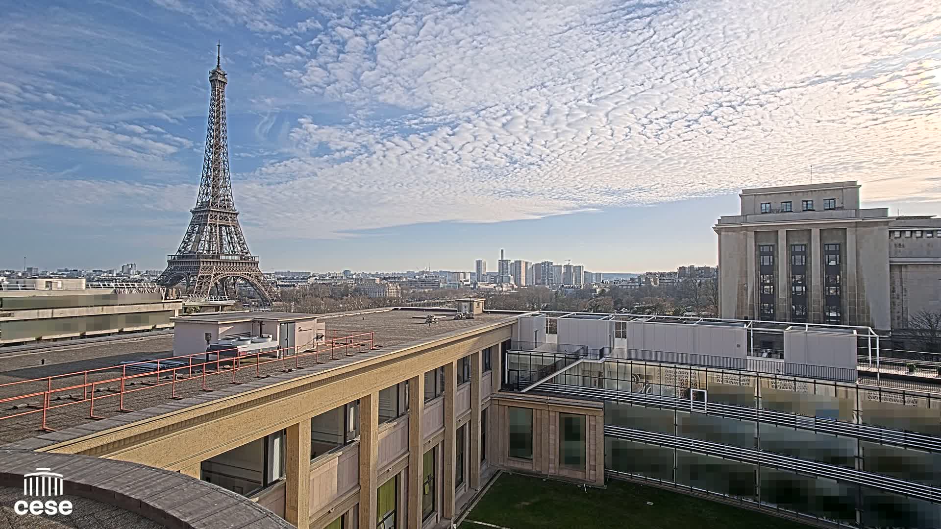 A view from a rooftop shows the Eiffel Tower in the distance under a partly cloudy sky, with other buildings and a cityscape visible in the background.