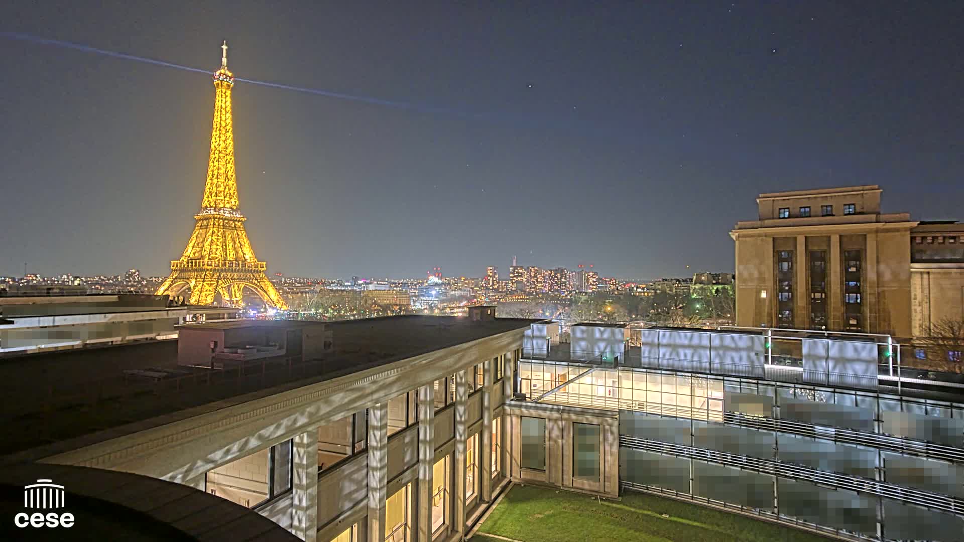 A nighttime view of the Eiffel Tower illuminated, a city skyline, and a courtyard with a building.