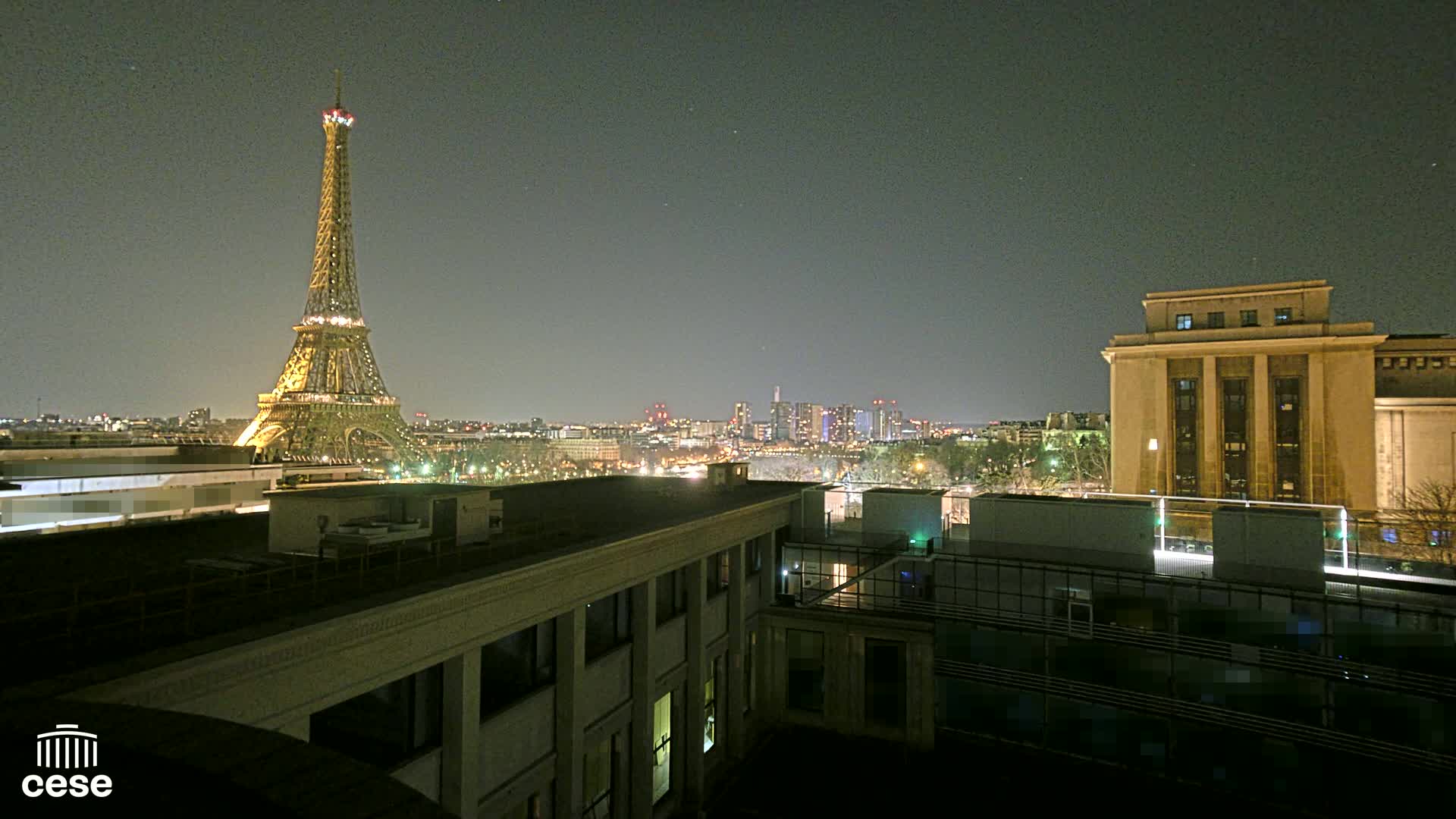 A nighttime view of the Eiffel Tower and a city skyline, seen from a rooftop overlooking another building.