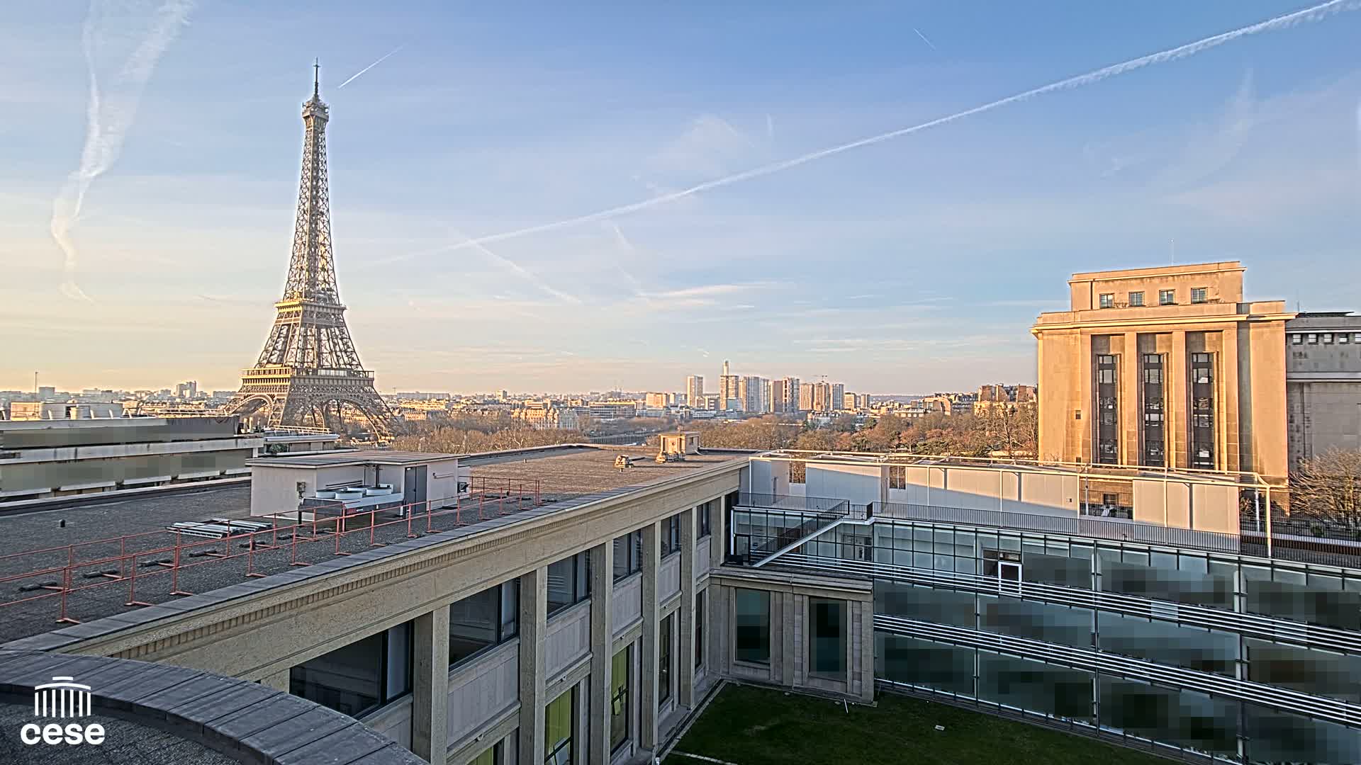 A rooftop view of Paris on a clear, sunny day shows the Eiffel Tower in the distance, alongside a large building and a cityscape.
