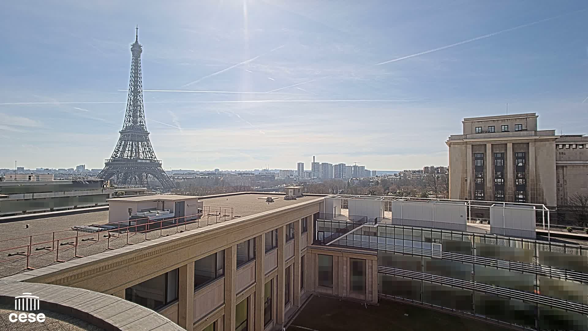 A sunny cityscape view shows the Eiffel Tower in the distance, beyond a flat rooftop with buildings and structures.