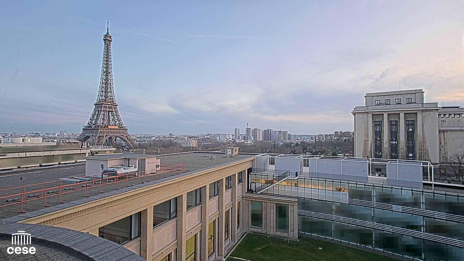An outdoor view shows the Eiffel Tower in the distance under a partly cloudy sky, alongside city buildings and a courtyard with modern architecture.