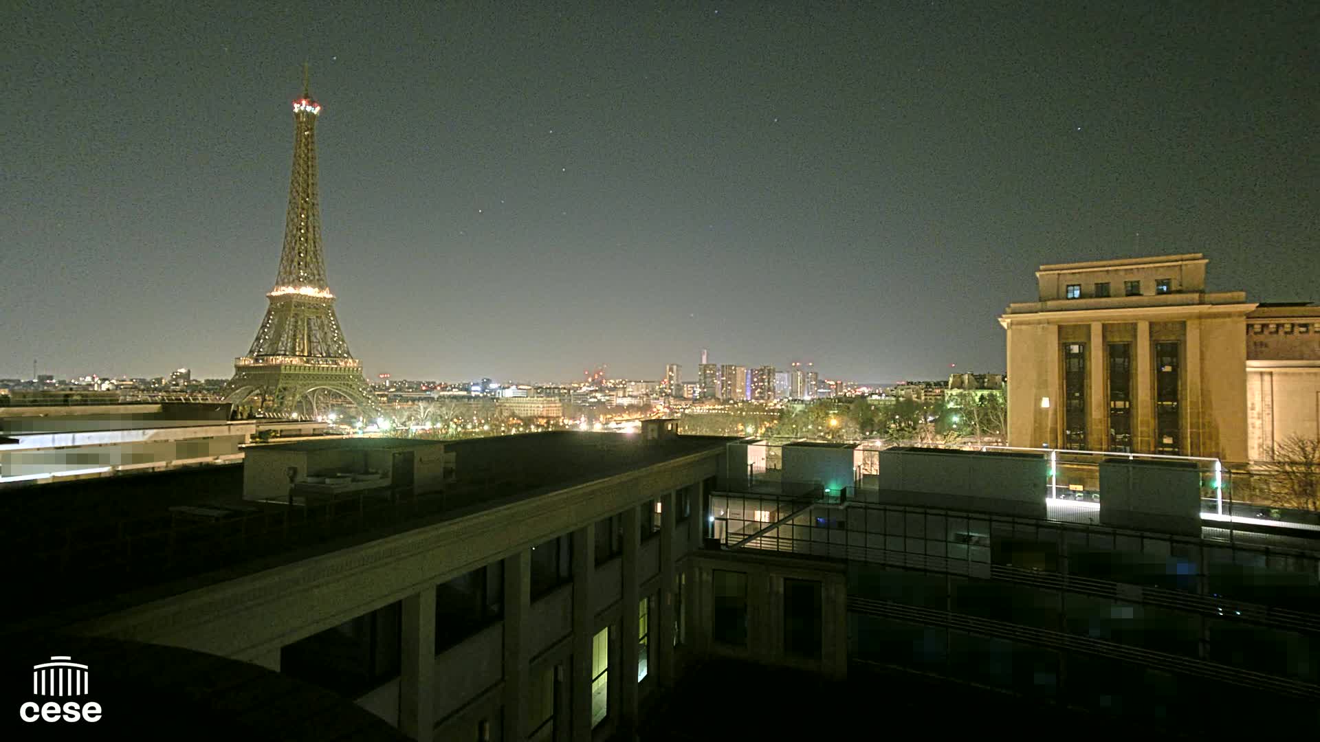 A nighttime cityscape view shows the Eiffel Tower and a city skyline, with a large building to the right and another in the foreground.