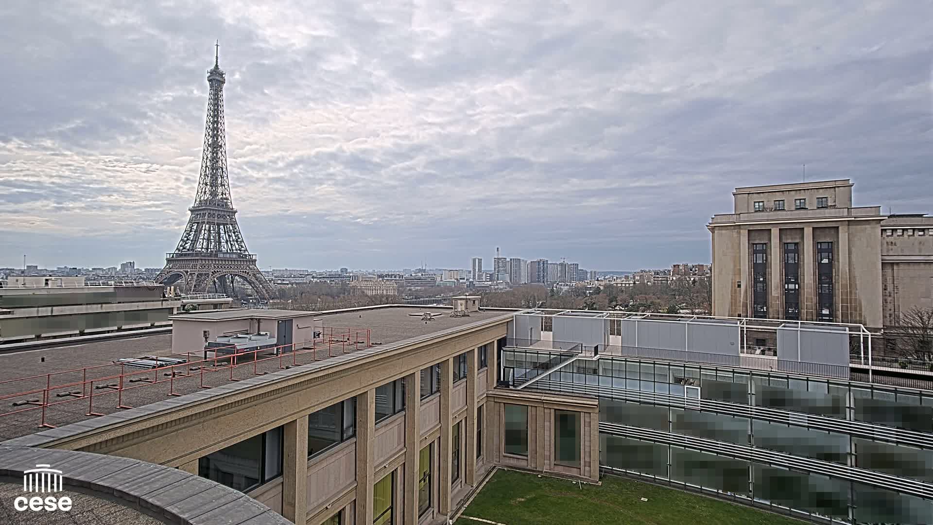 A view from a rooftop shows the Eiffel Tower in the distance under a cloudy sky, with modern and classical buildings in the foreground.