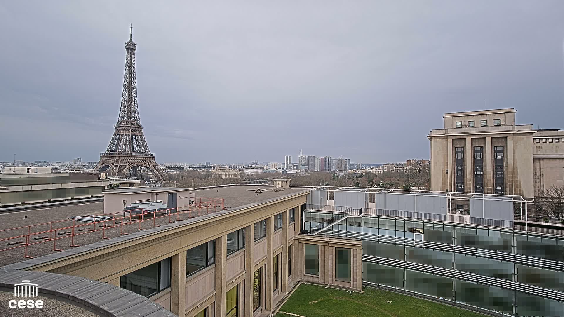 A rooftop view of Parisian architecture, including the Eiffel Tower in the distance under a cloudy sky, shows a tan building with a flat roof and another building with a classical facade.