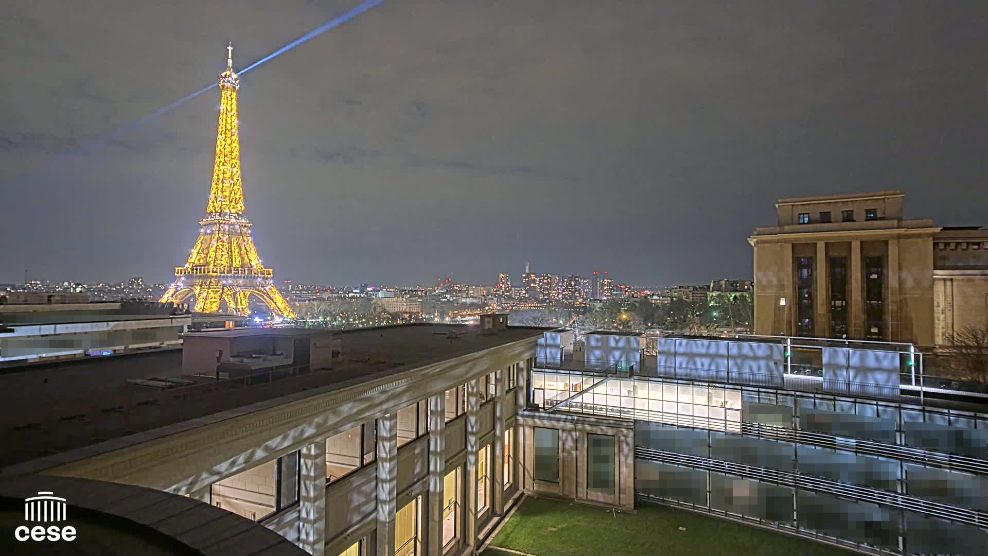 A nighttime view of the Eiffel Tower illuminated, along with a city skyline and adjacent buildings, under a dark, cloudy sky.