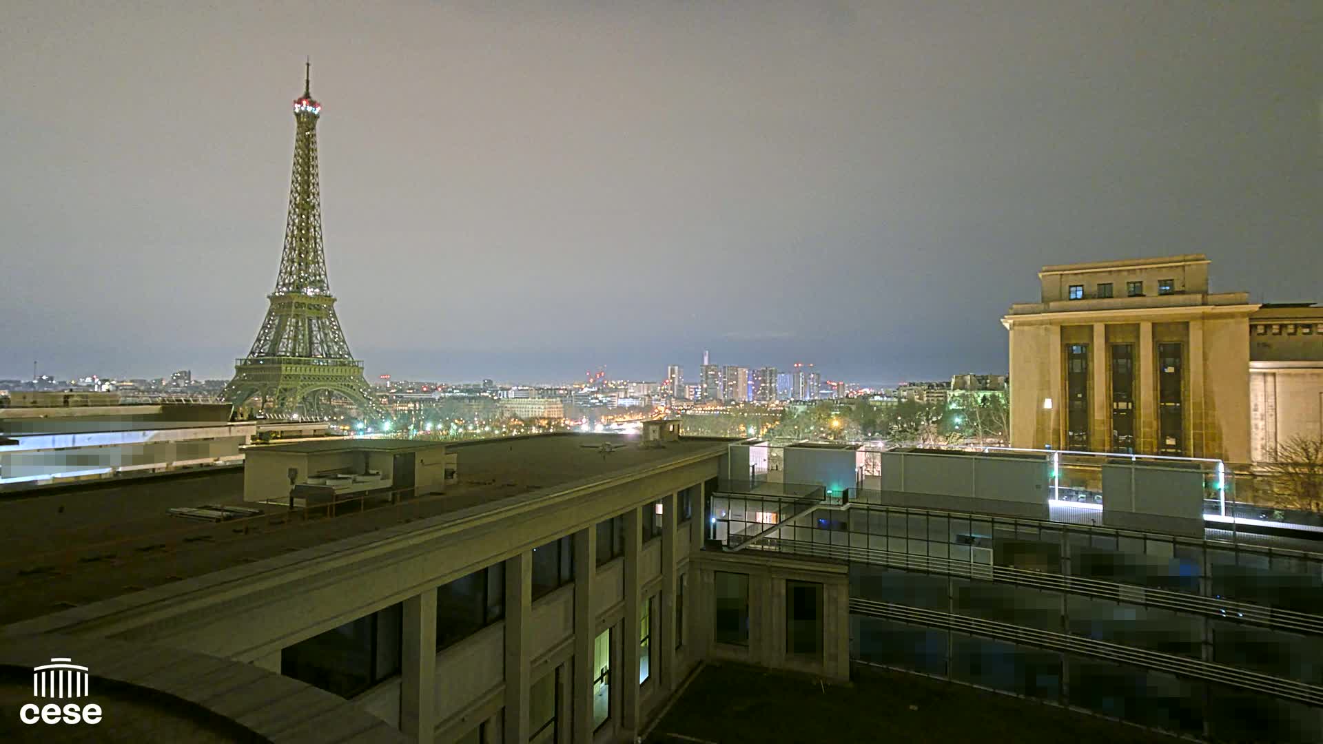 A nighttime view of the Eiffel Tower and a city skyline from a rooftop, under a hazy sky.