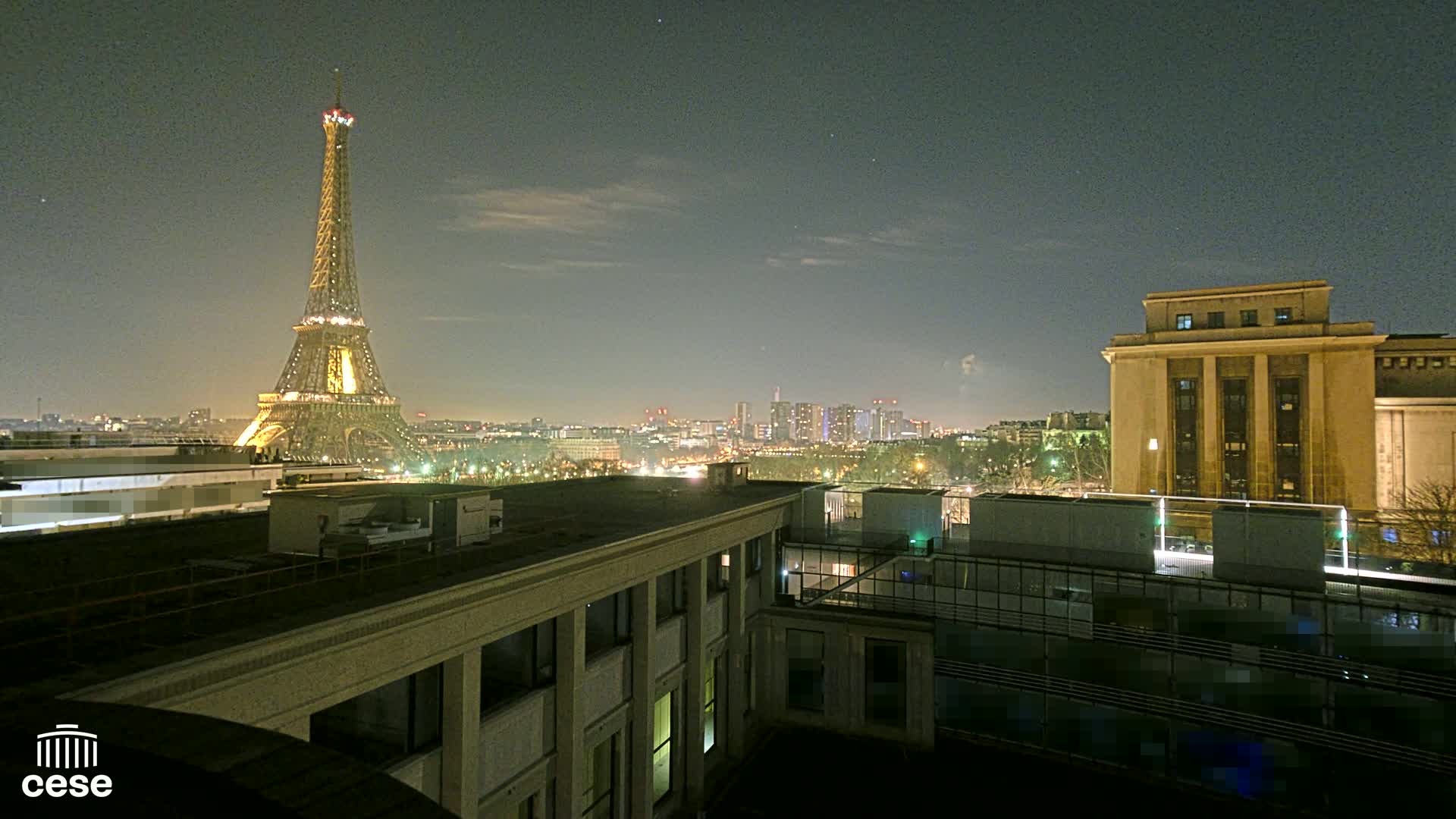 A nighttime view of the Eiffel Tower illuminated, a city skyline, and a large building, under a mostly clear sky.