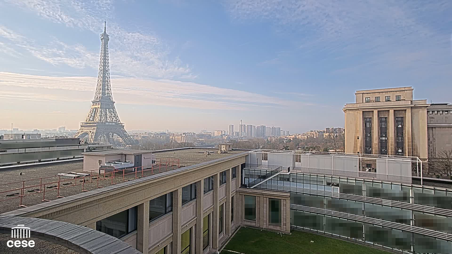 A view from a rooftop shows the Eiffel Tower in the distance under a mostly clear, bright sky, with a cityscape and modern buildings in the foreground.