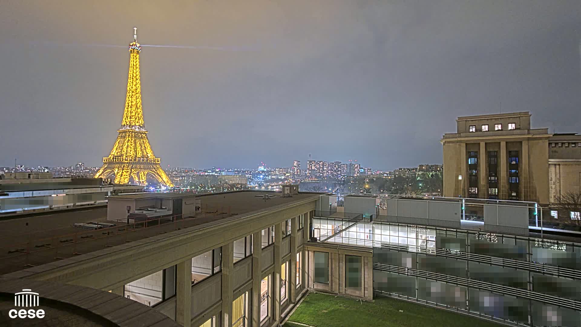 A nighttime view of the Eiffel Tower illuminated in gold, seen from a rooftop overlooking a city with overcast skies.