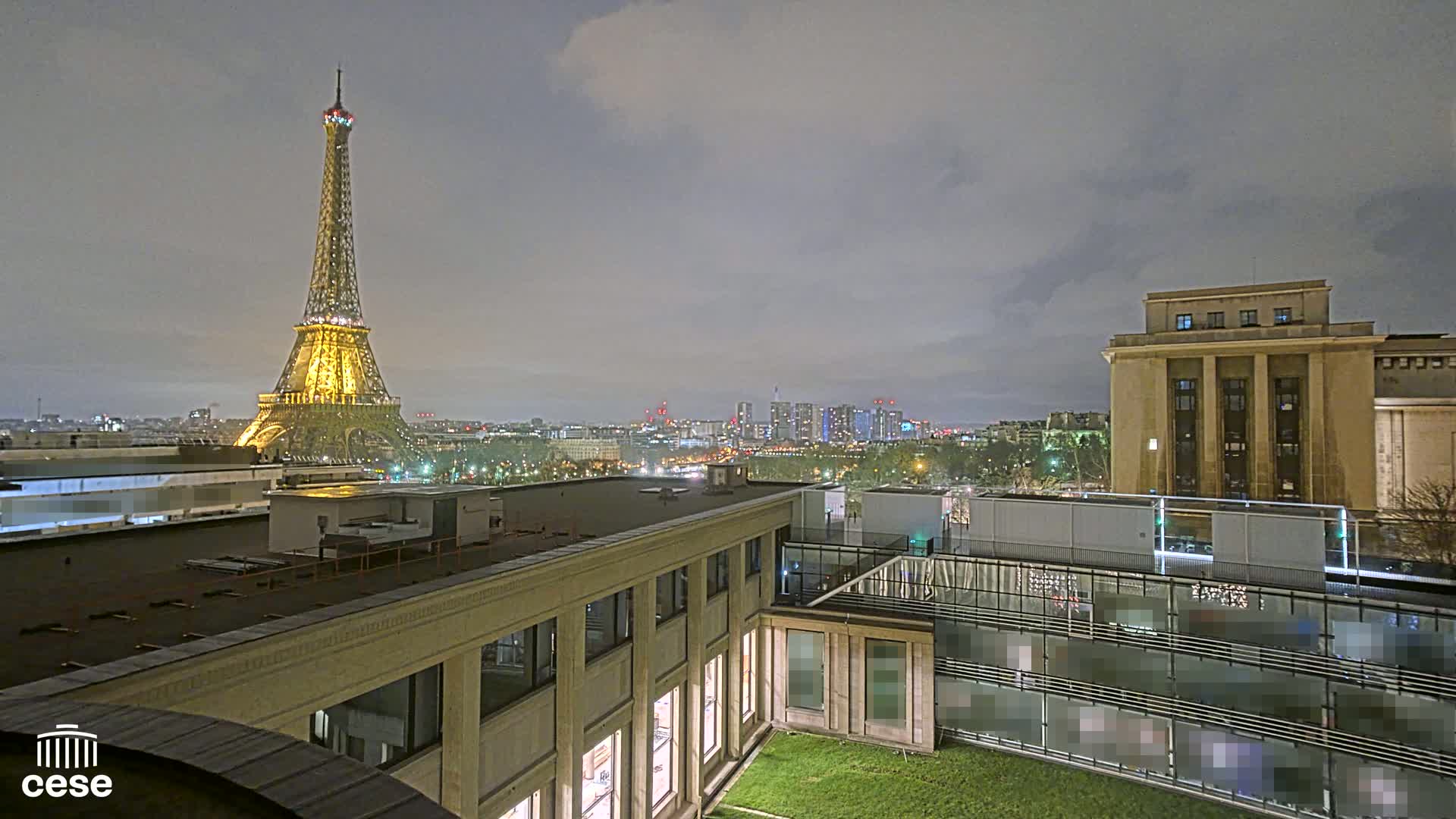 A nighttime view of the Eiffel Tower illuminated against a cloudy sky, overlooking a city skyline and rooftops, including a large building with a flat roof.