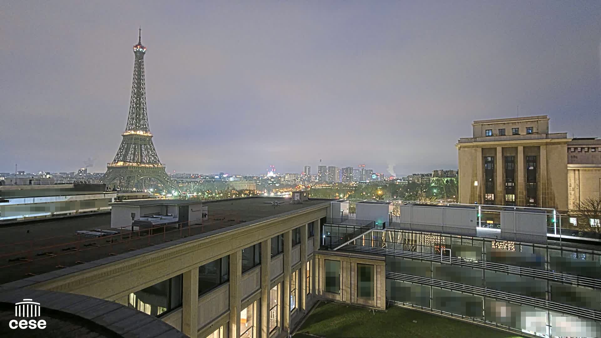 A nighttime view of the Eiffel Tower and a cityscape, seen from a rooftop with other buildings nearby under an overcast sky.
