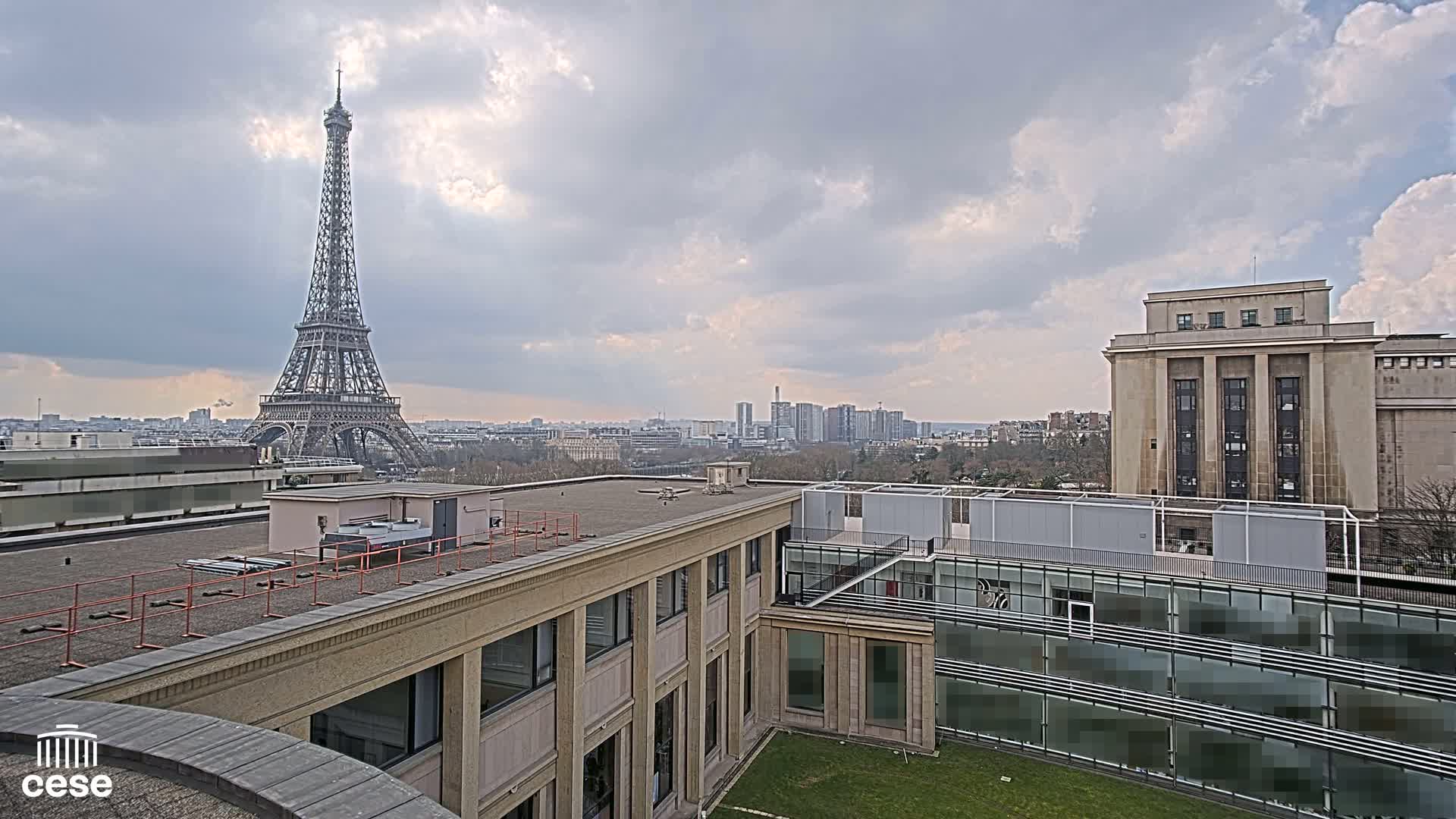 A view from a rooftop shows the Eiffel Tower in the distance under a cloudy sky, with other buildings and a grassy courtyard in the foreground.